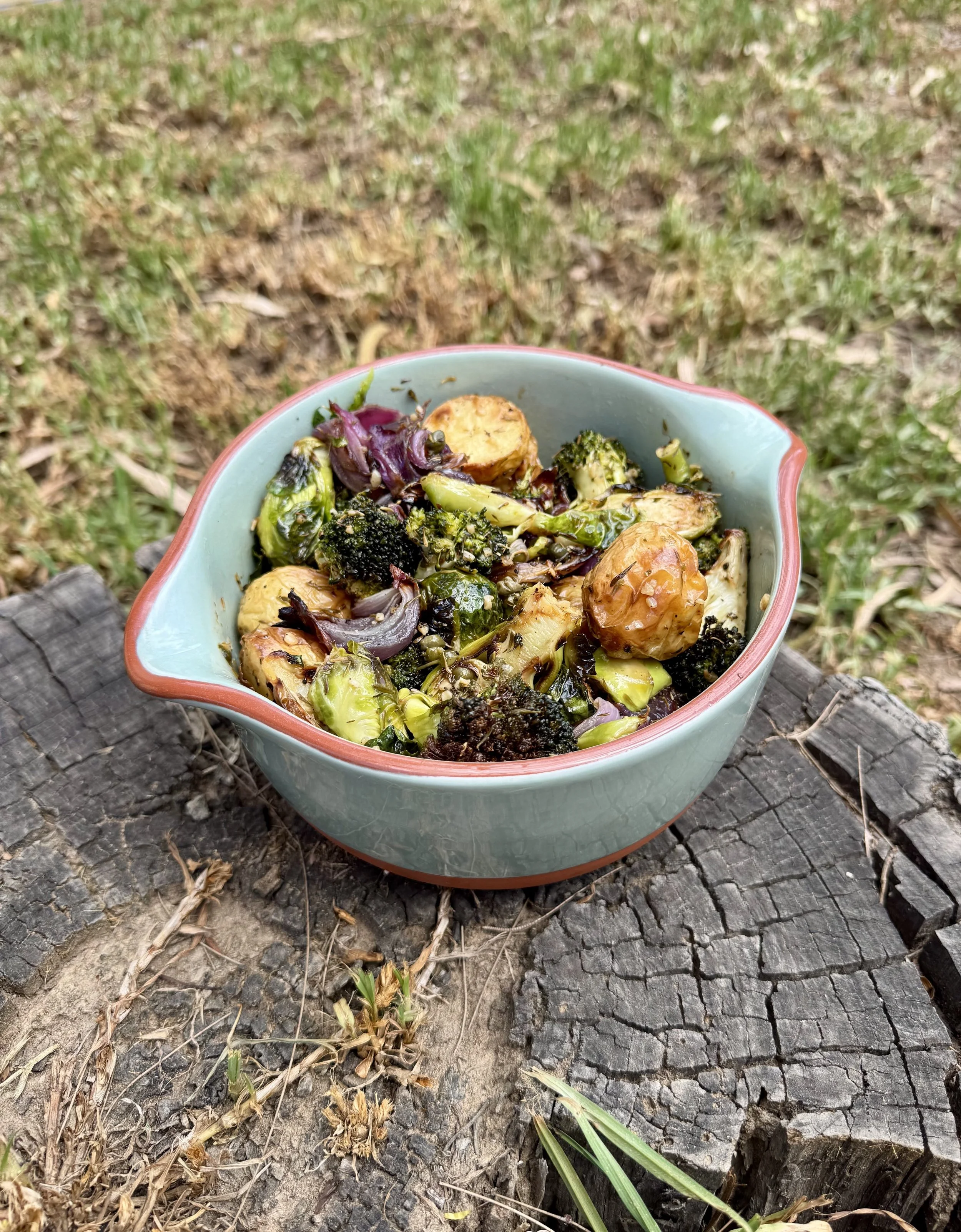 A ceramic bowl filled with roasted vegetables, including broccoli, Brussels sprouts, and purple onions, sits on a weathered wood surface outdoors with grass and dirt in the background.