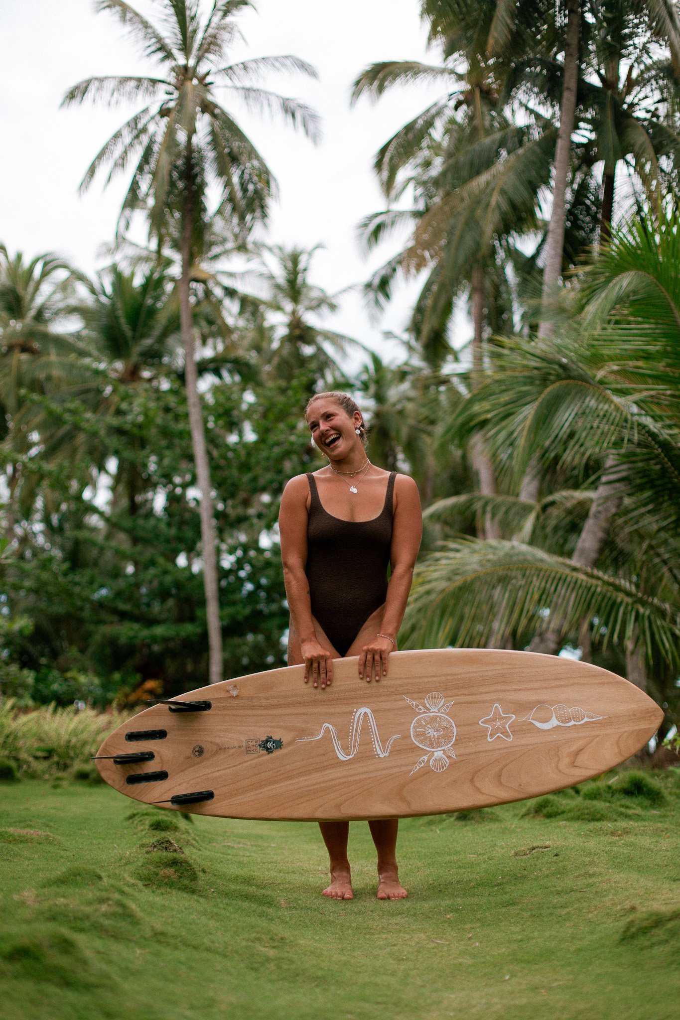 A woman standing on grass, holding a large surfboard with white marine-themed drawings, surrounded by tall palm trees.