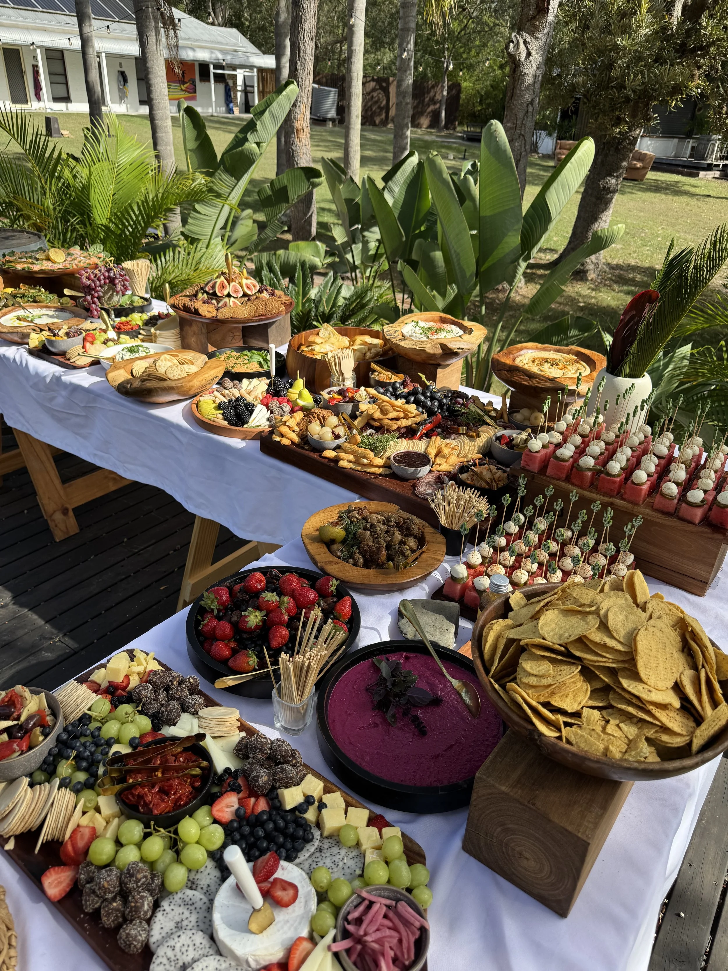 Outdoor buffet table filled with various fruits, cheeses, dips, crackers, vegetables, and appetizers set on a white tablecloth with surrounding greenery and trees in the background.