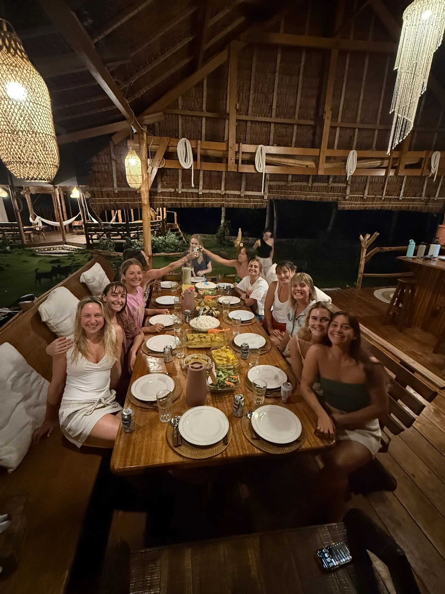 Group of young women sitting around a large wooden table outdoors at night, smiling and enjoying a meal with various dishes, drinks, and a shared toast, under a rustic thatched roof structure decorated with hanging lanterns.