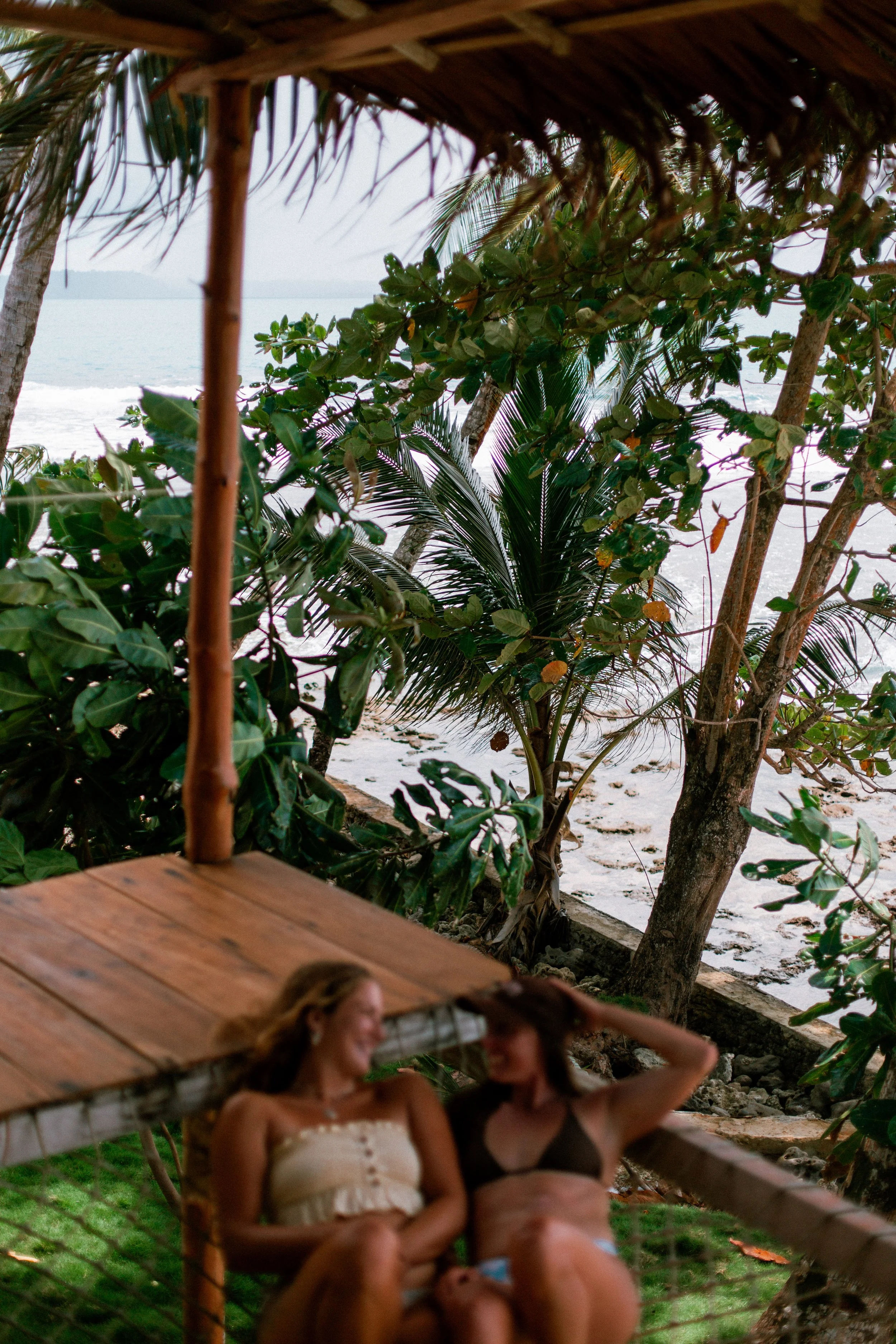 Two women relaxing on a porch, with tropical trees, beach, and ocean in the background.
