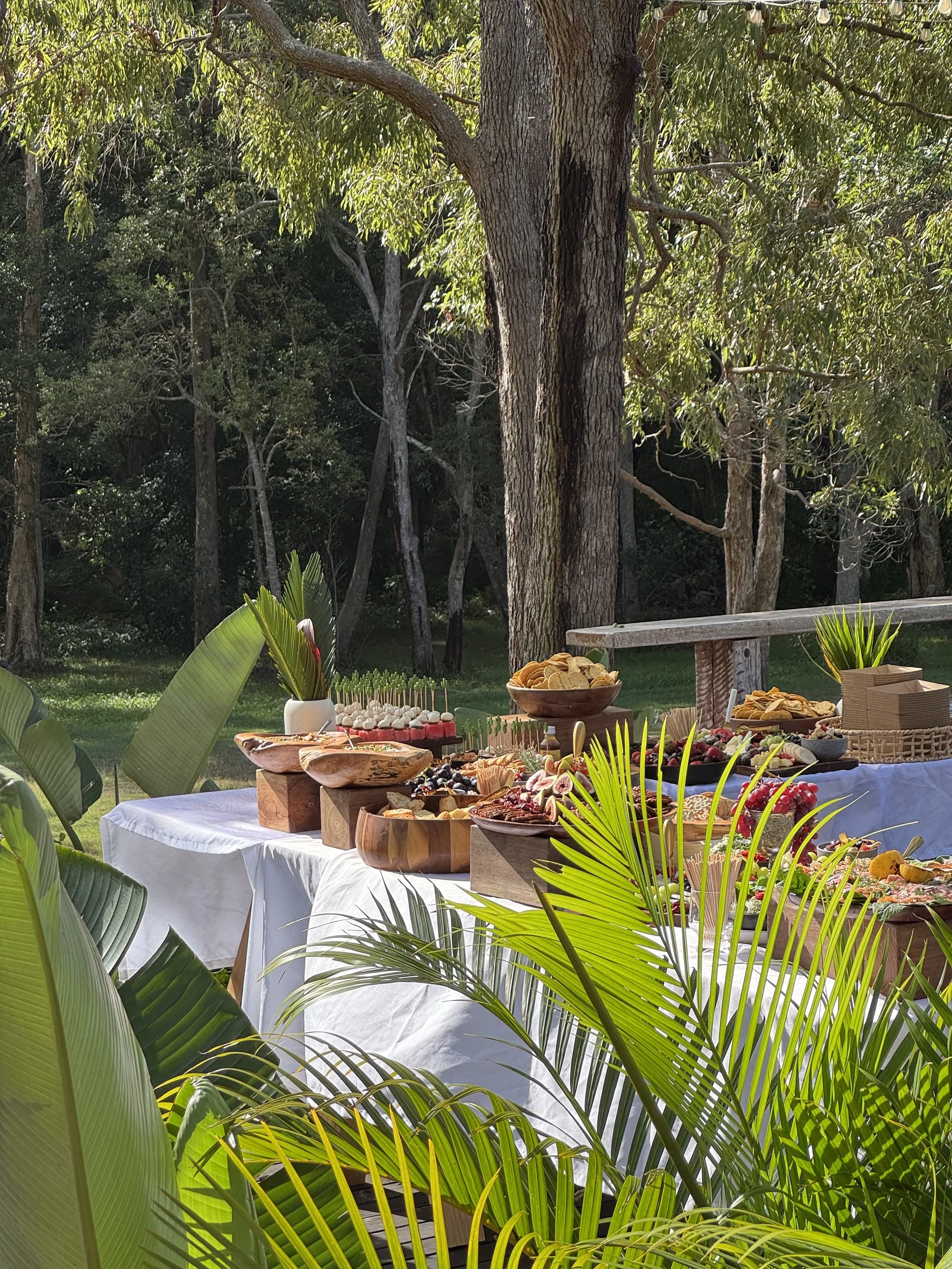 Outdoor buffet table with various dishes, surrounded by green tropical plants and trees.