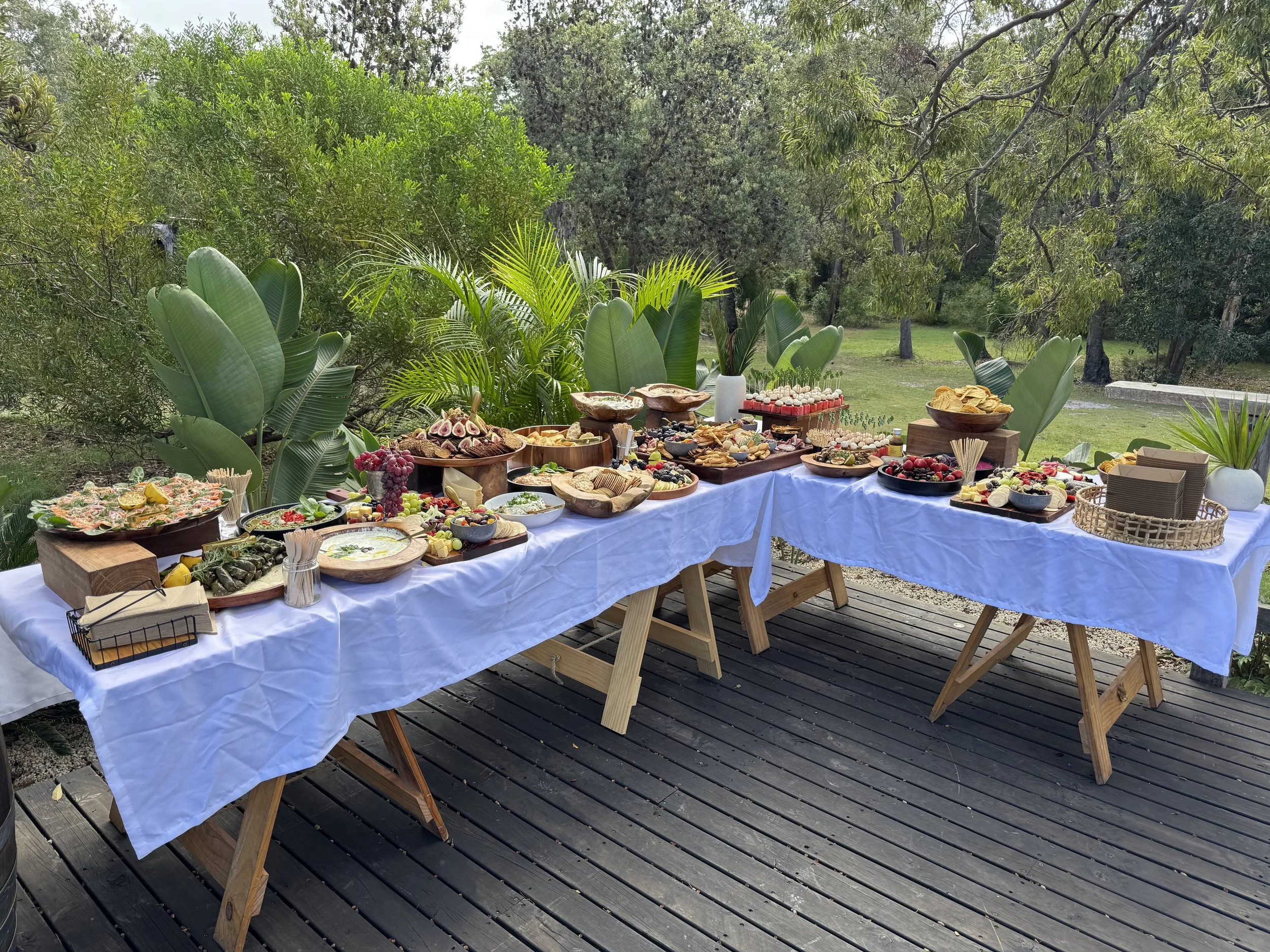 Outdoor table set up for a meal with a white tablecloth, surrounded by lush green trees and plants.