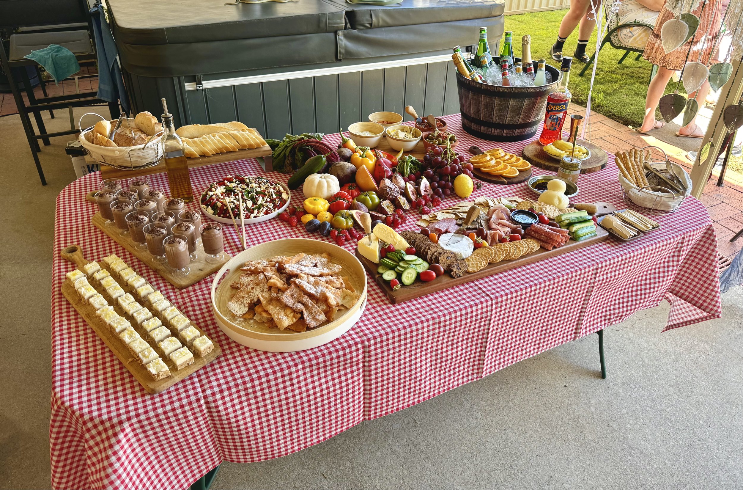 A picnic table with a red and white checkered tablecloth holding various foods including desserts, fruits, salads, bread, and drinks. In the background, there is a hot tub and some people standing outside.