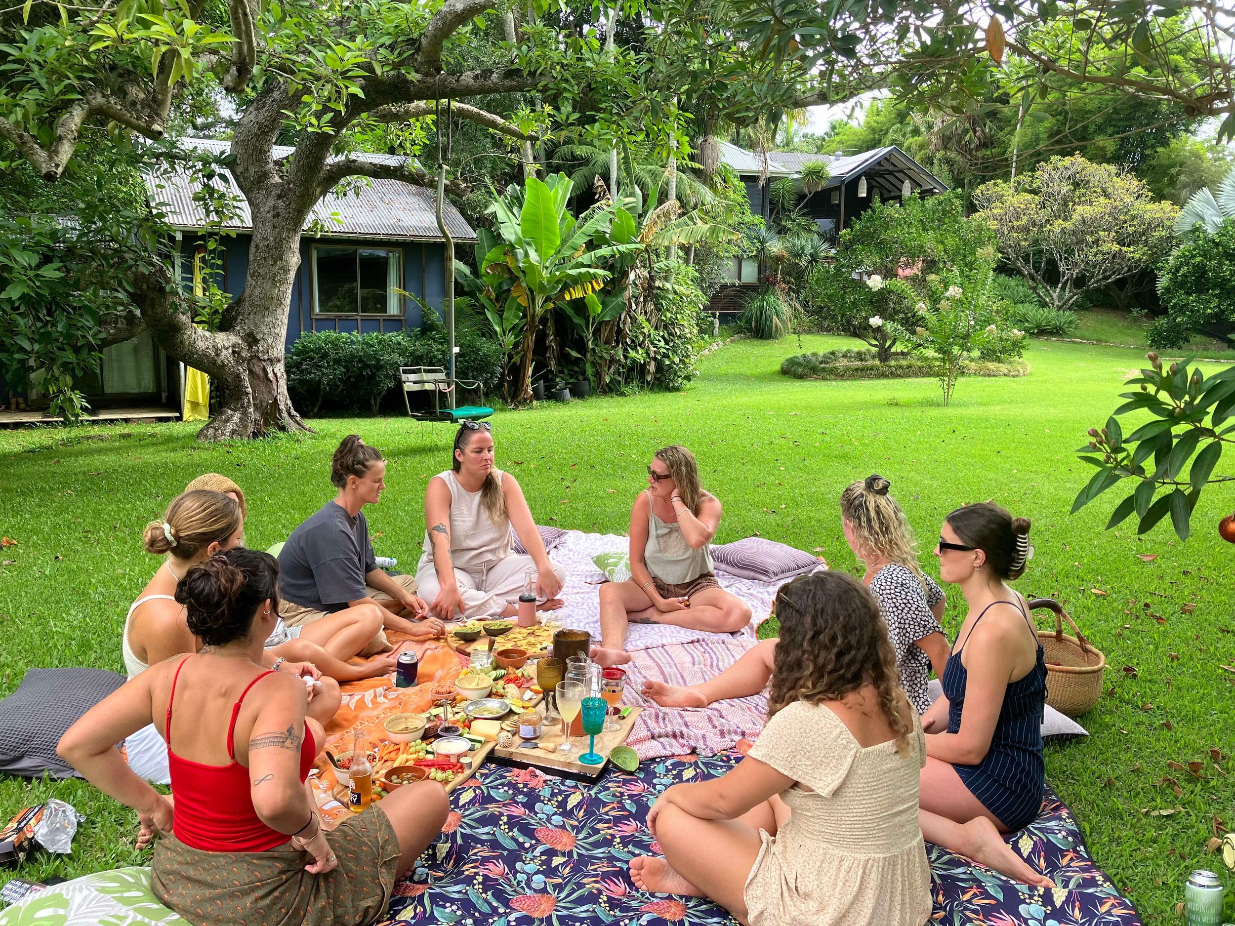 A group of people sitting cross-legged on colorful blankets and cushions on a green lawn, enjoying a meal outdoors under a large tree with green foliage. There are tables filled with various foods, drinks, and utensils. In the background, there are houses with metal roofs surrounded by lush tropical plants.