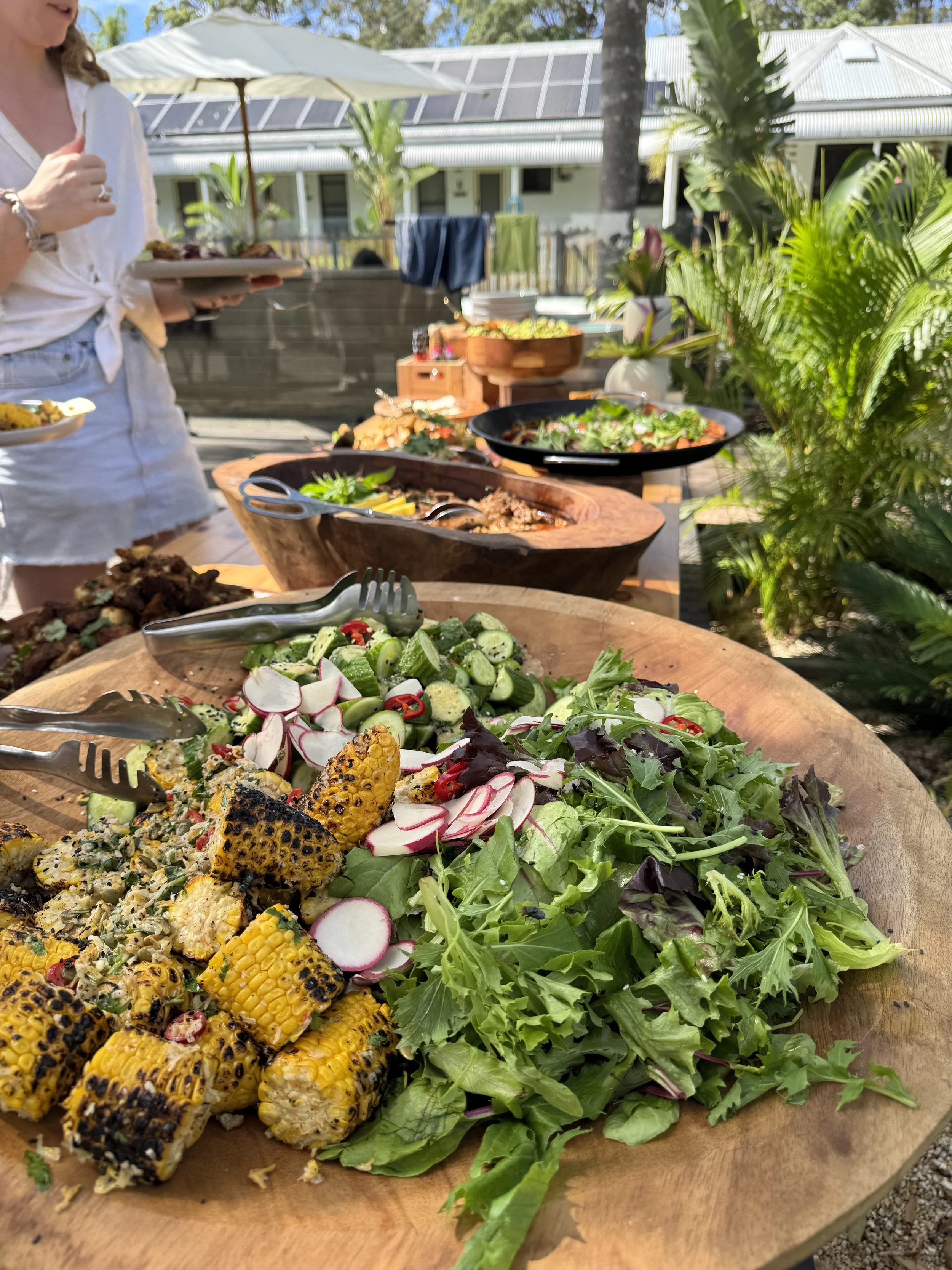 A wooden platter with grilled corn, radish slices, and mixed greens, set outdoors on a table, with a person holding a plate of food and a backyard with plants and buildings in the background.