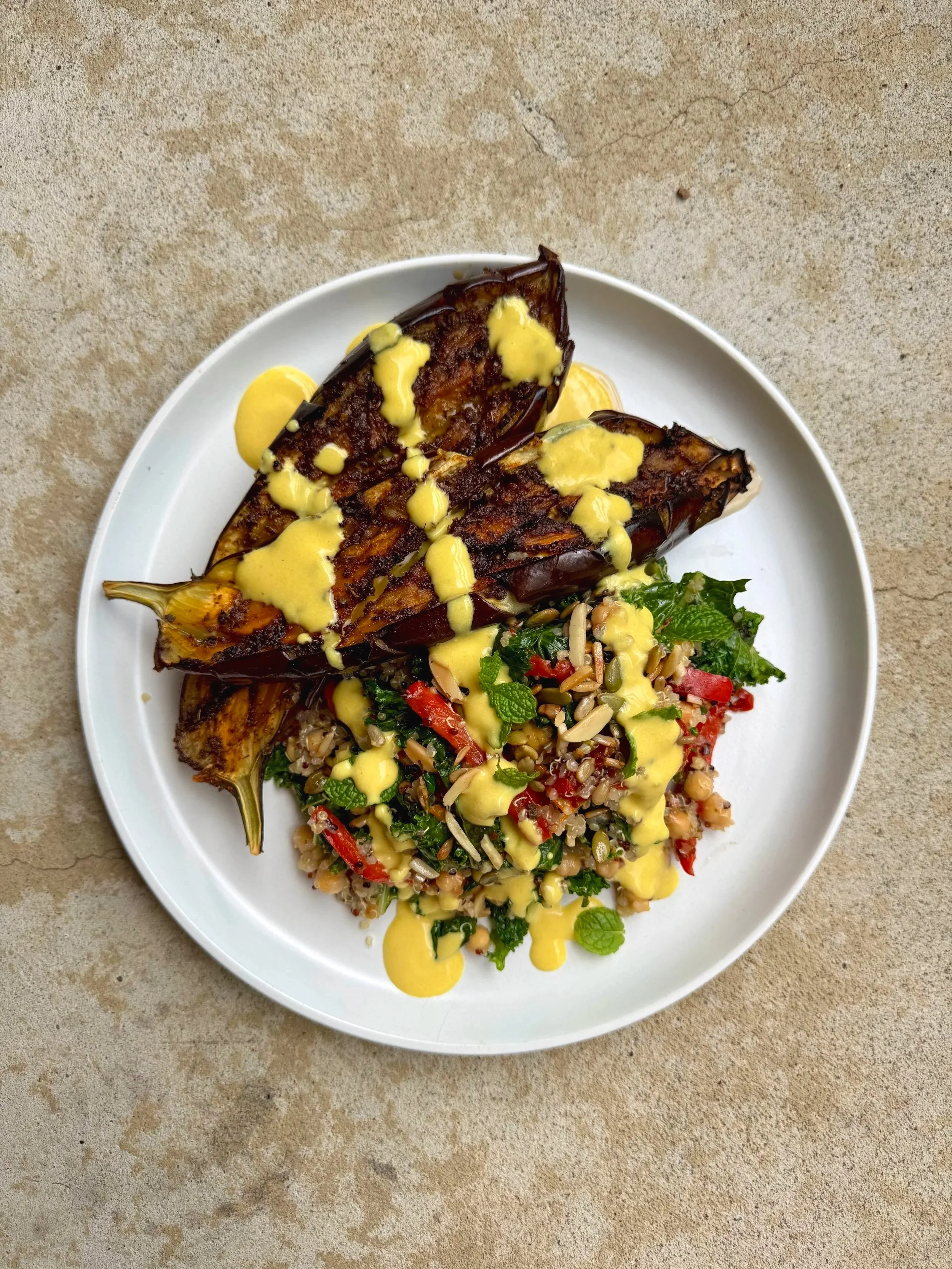 Plate of grilled fish topped with yellow sauce, alongside a vegetable and grain salad with cherry tomatoes, greens, herbs, and seeds on a neutral surface.