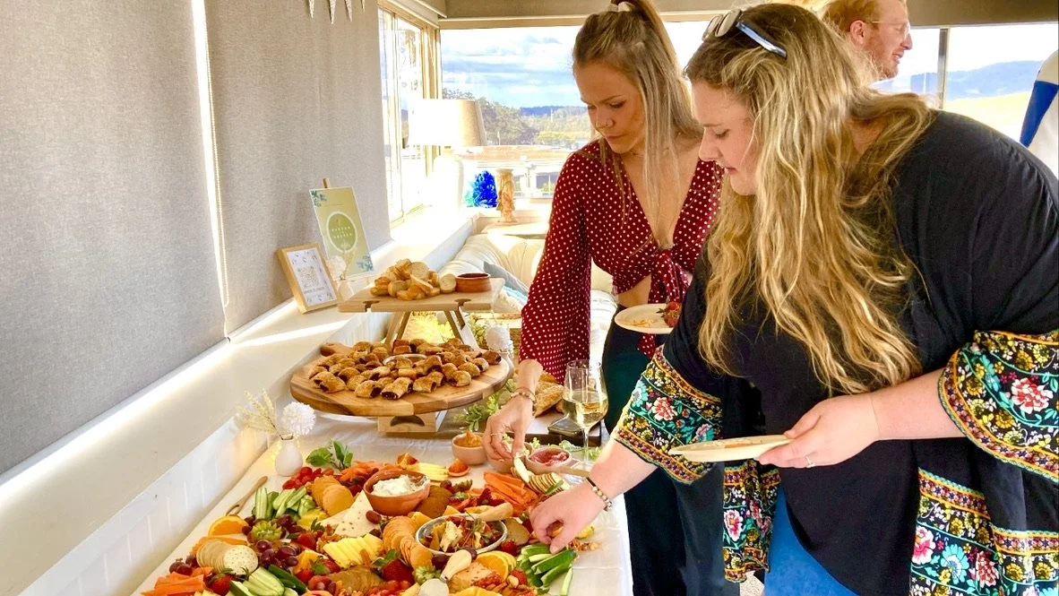 Two women reaching for food on a buffet table with various dishes, including fruit, cheeses, and snacks, in a bright room with windows in the background.