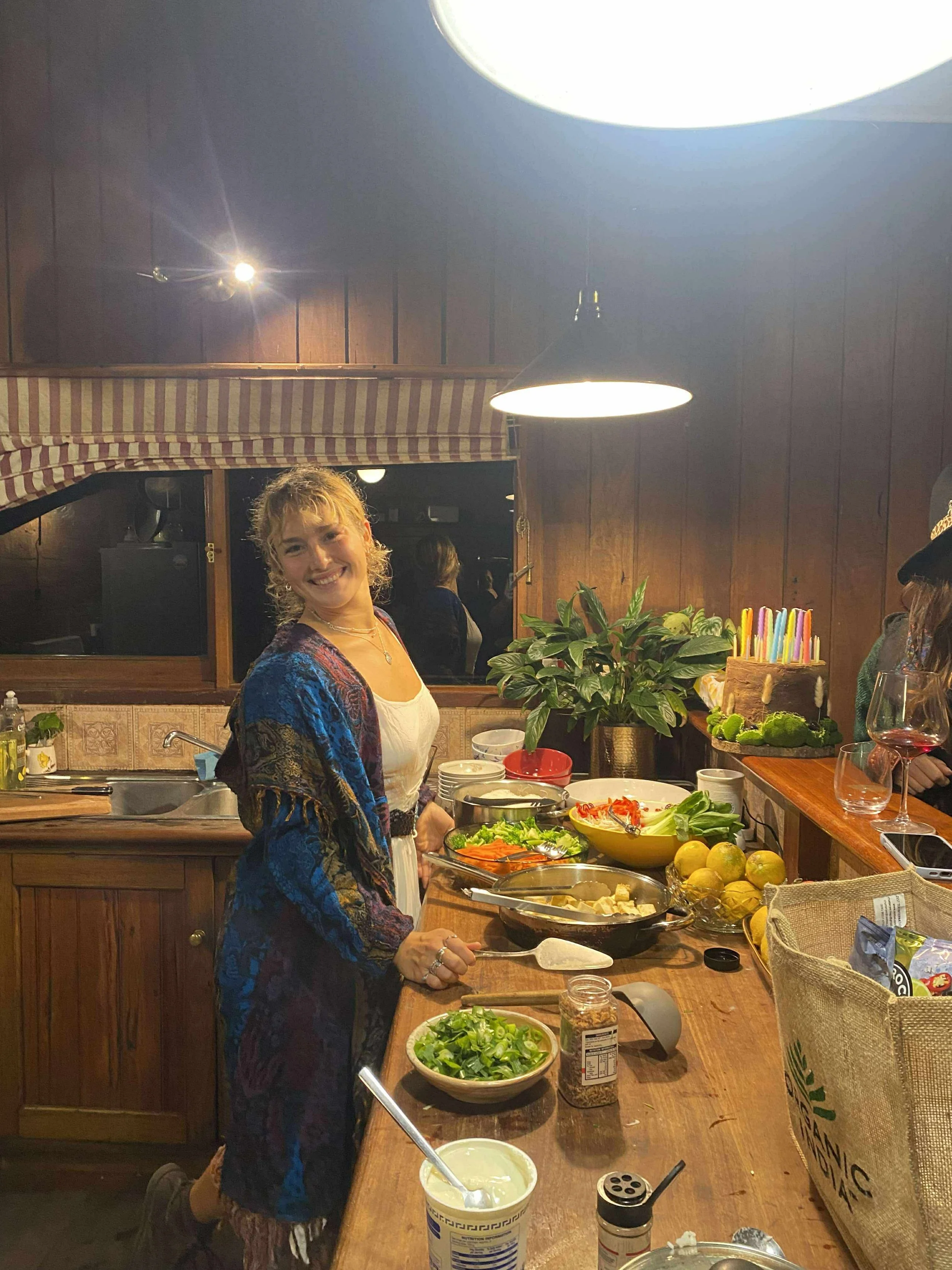 A woman preparing food in a kitchen with a variety of vegetables and ingredients on the counter, smiling at the camera.