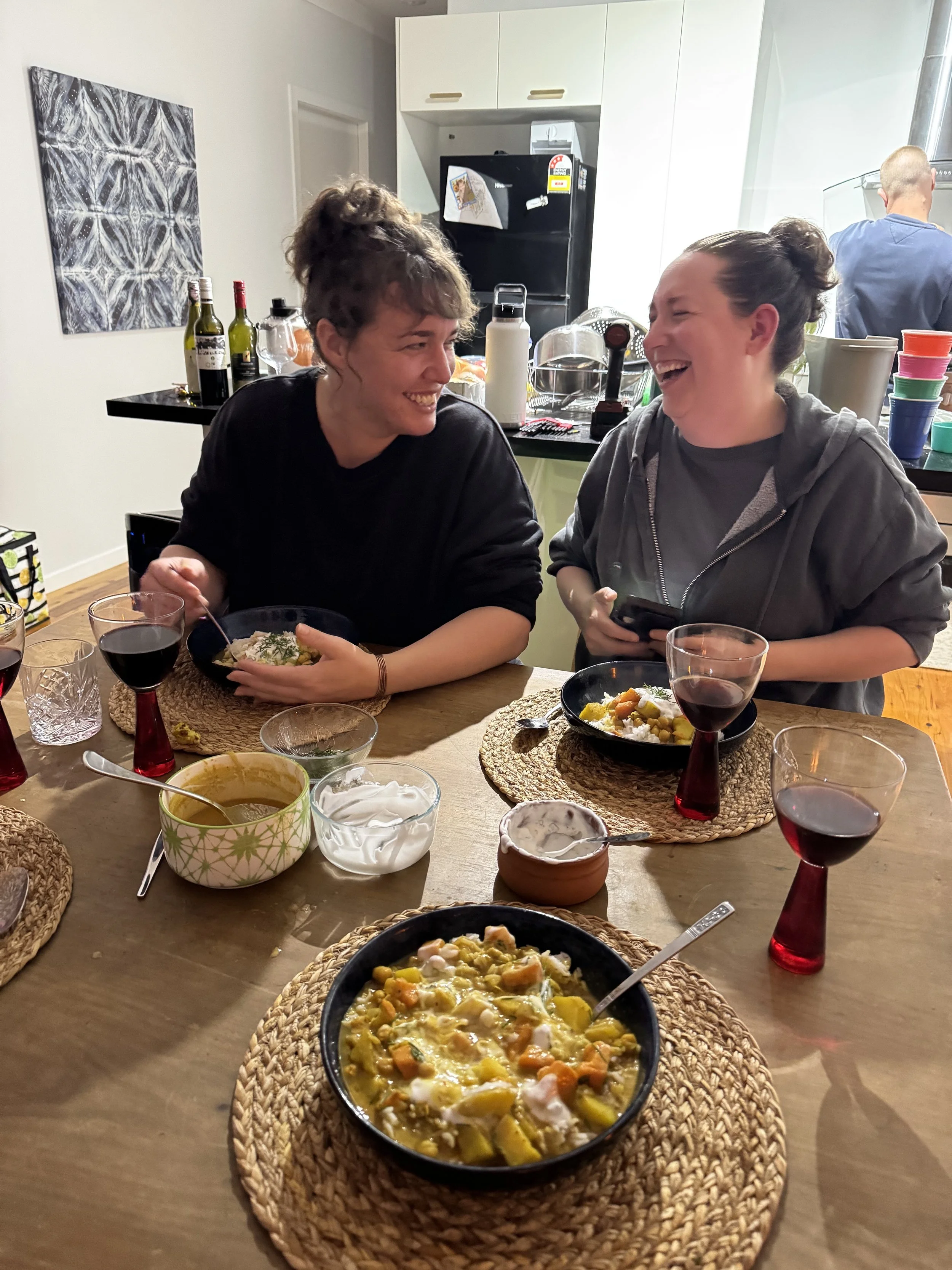 People sitting at a table with bowls of food, drinks, and various condiments, laughing and enjoying each other's company in a cozy, well-lit room.