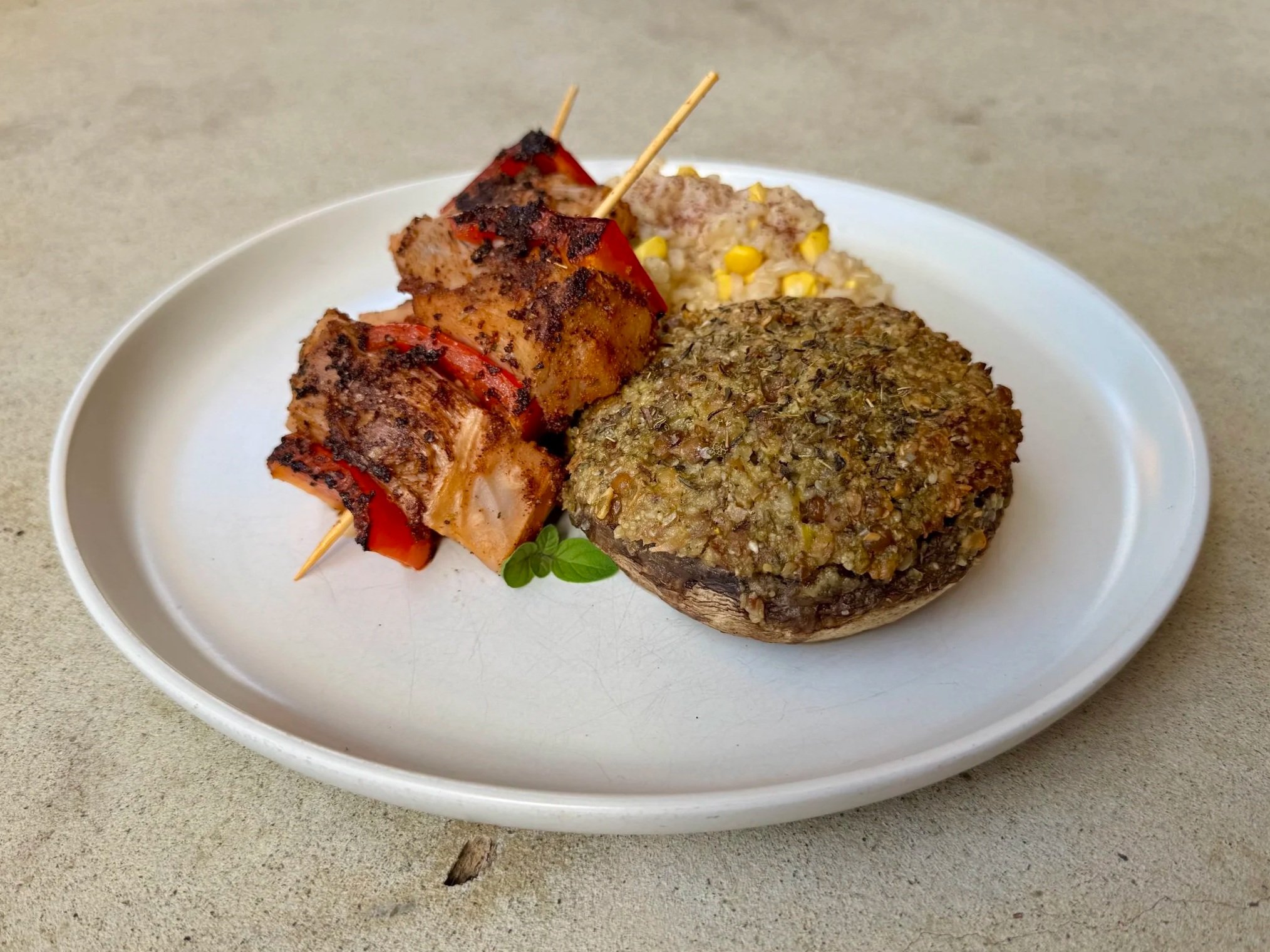 A white plate with a skewer of grilled meat and red bell peppers, a portion of cooked rice with corn, and a large cooked mushroom with herbs, all on a light-colored surface.