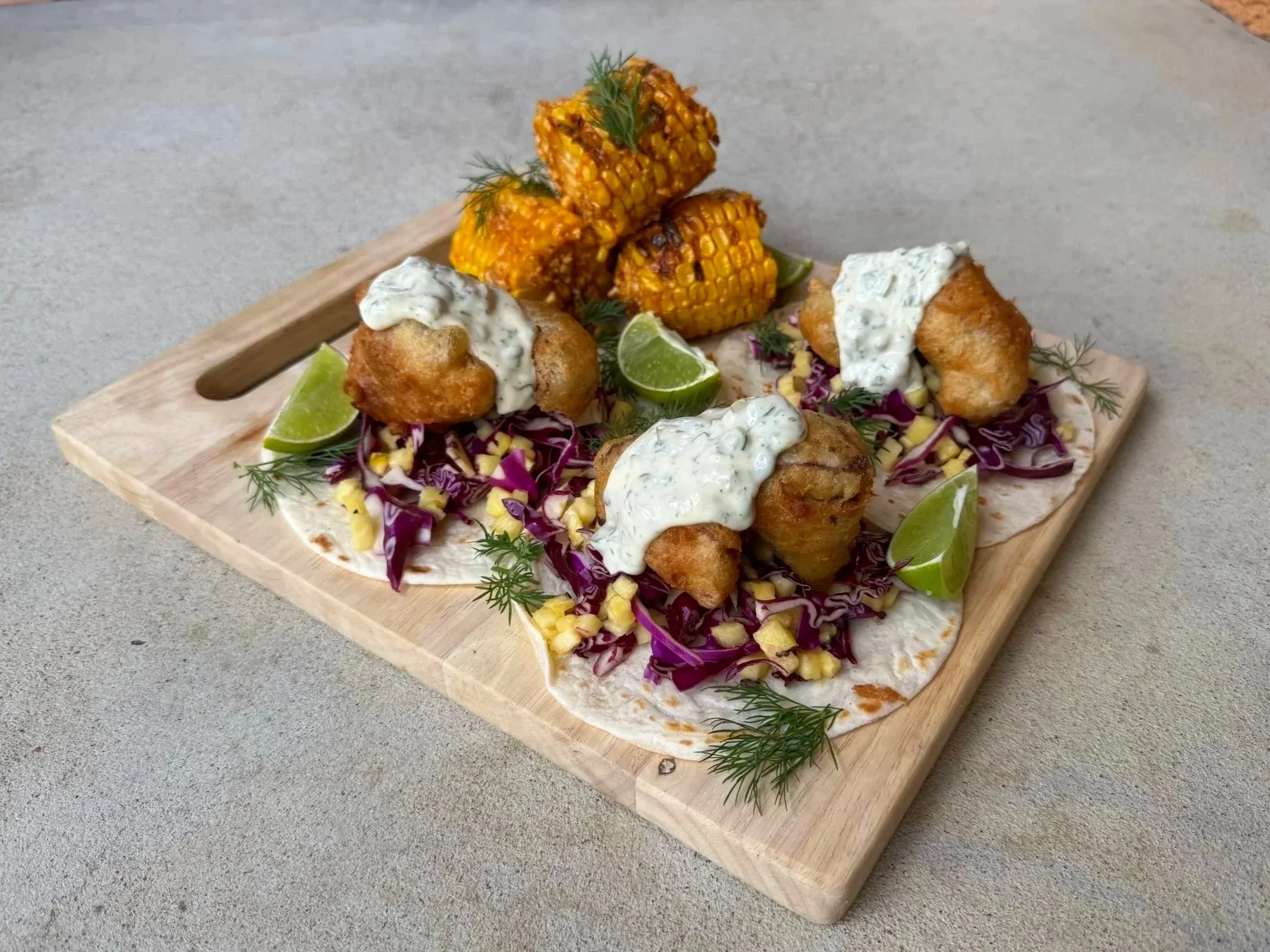 A wooden platter with fried fish fillets topped with creamy white sauce, lime wedges, shredded purple cabbage and yellow corn, garnished with dill. In the background, grilled corn on the cob on top of cabbage and dill, on a gray surface.