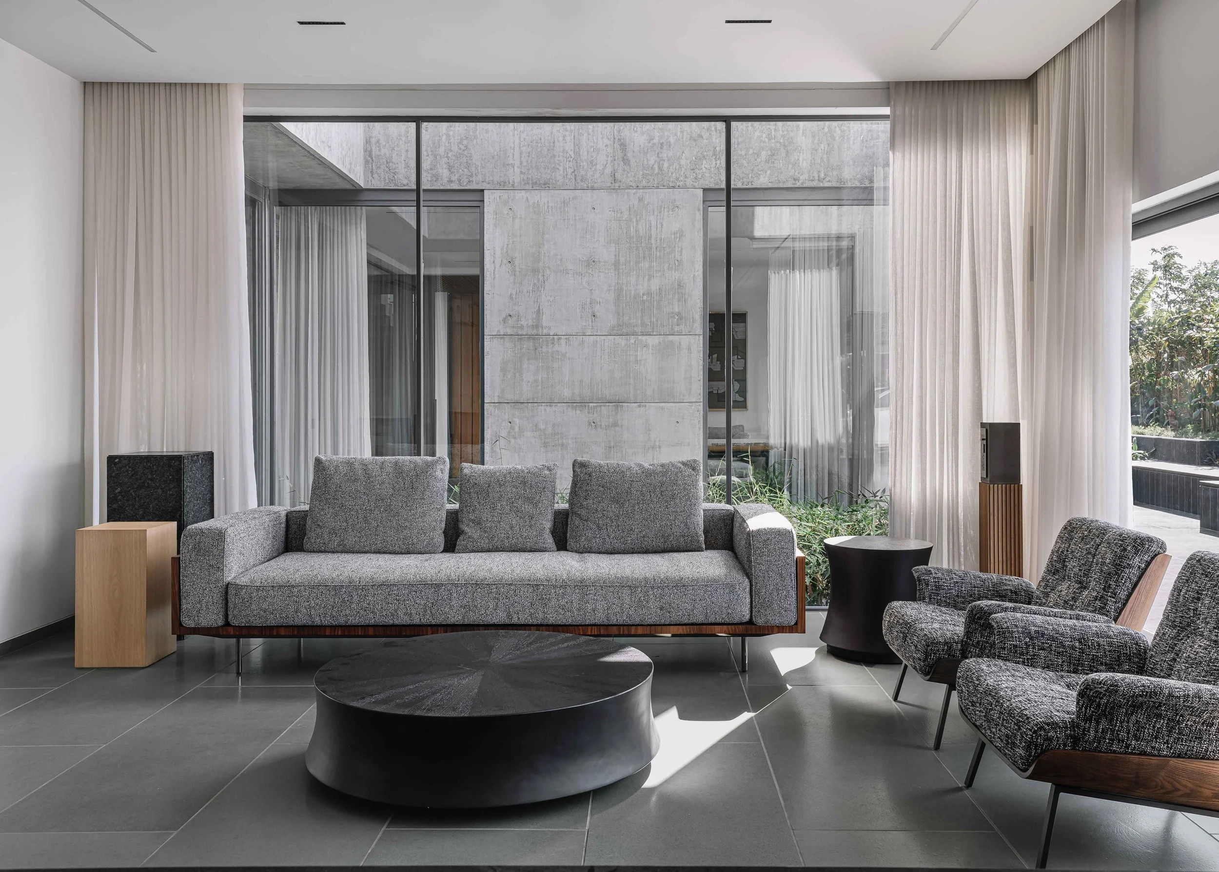 Modern living room with grey sofa, patterned armchairs, black coffee table, large windows with white curtains, and minimalist decor.