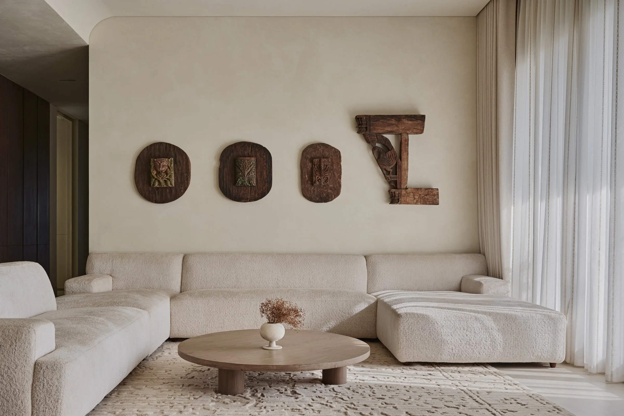 Living room with off-white sectional sofa, wooden coffee table with a white vase and dried flowers, neutral-colored rug, white curtains, and wall decorations made of dark wood carved plaques and a letter 'T' sculpture.