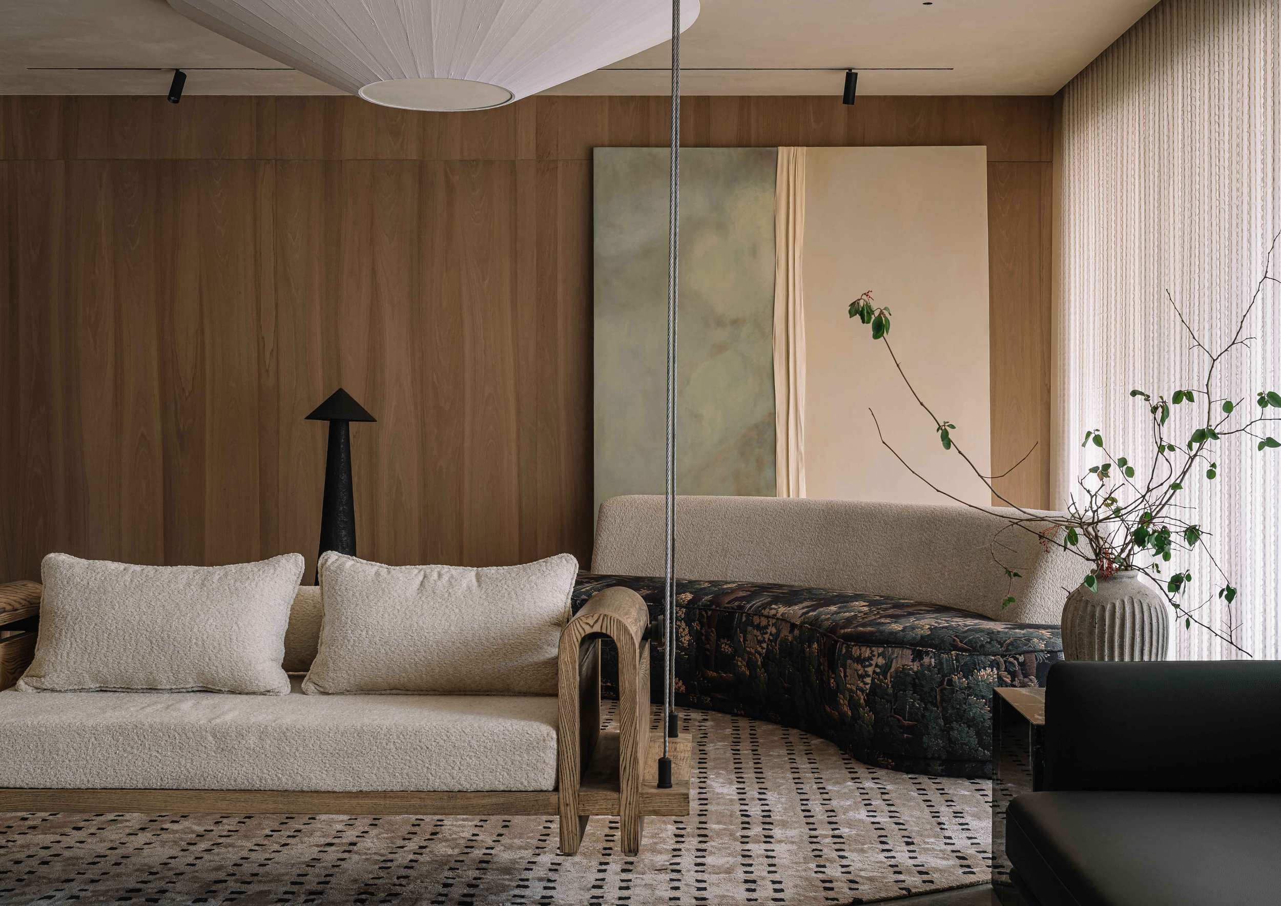 Modern living room with beige textured sofas, a black lamp, a wooden wall, an abstract painting, a vase with branches, and a patterned rug.