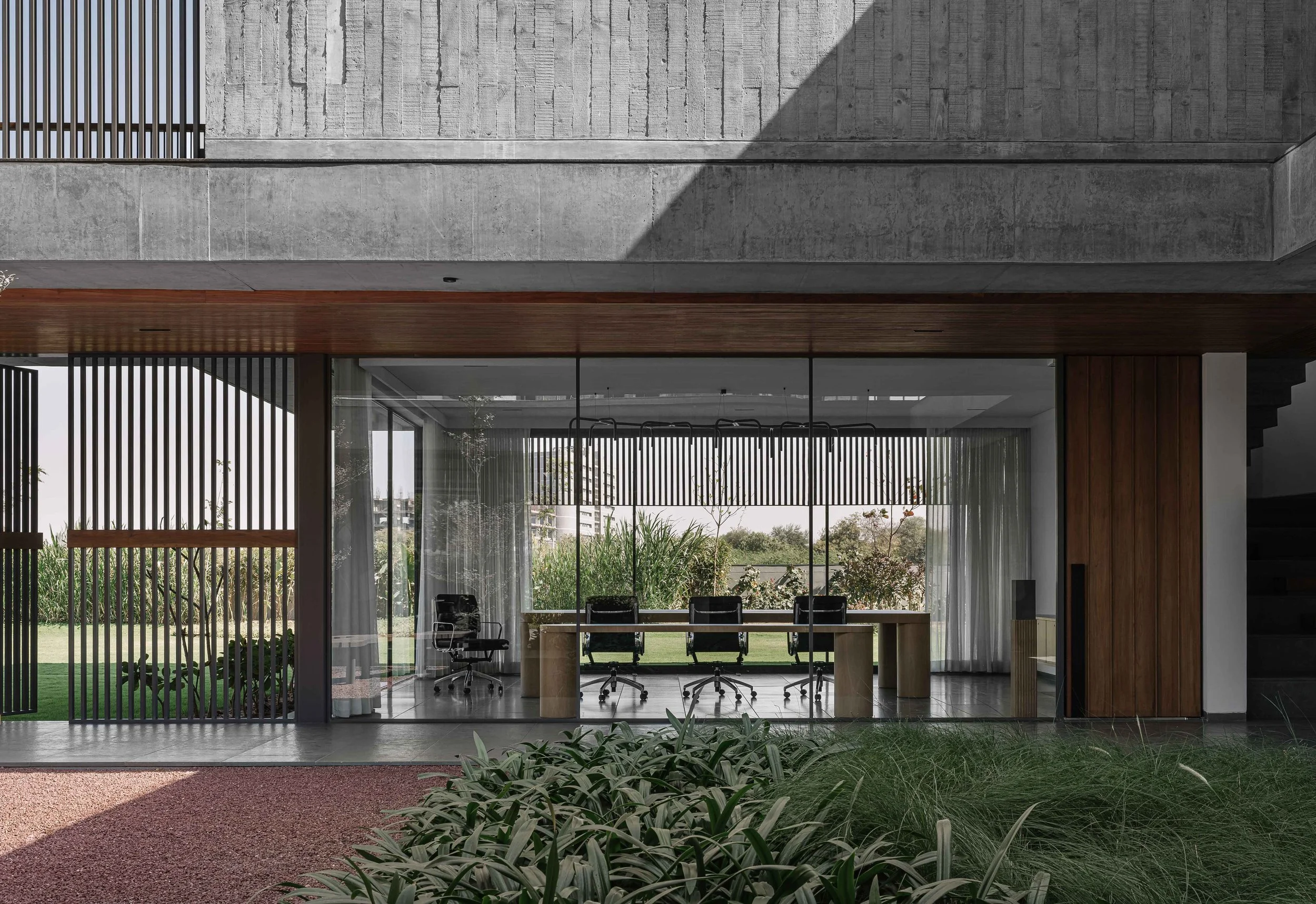 Modern office space with glass walls, a wooden ceiling, and three black chairs around a large desk. Outside, greenery and trees are visible through the windows.