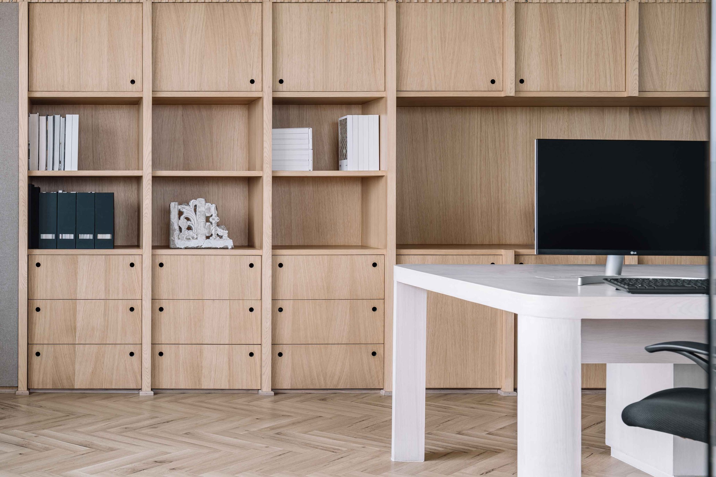 Office space with a wooden bookshelf filled with books and decorative items, and a white desk with a computer monitor and keyboard.