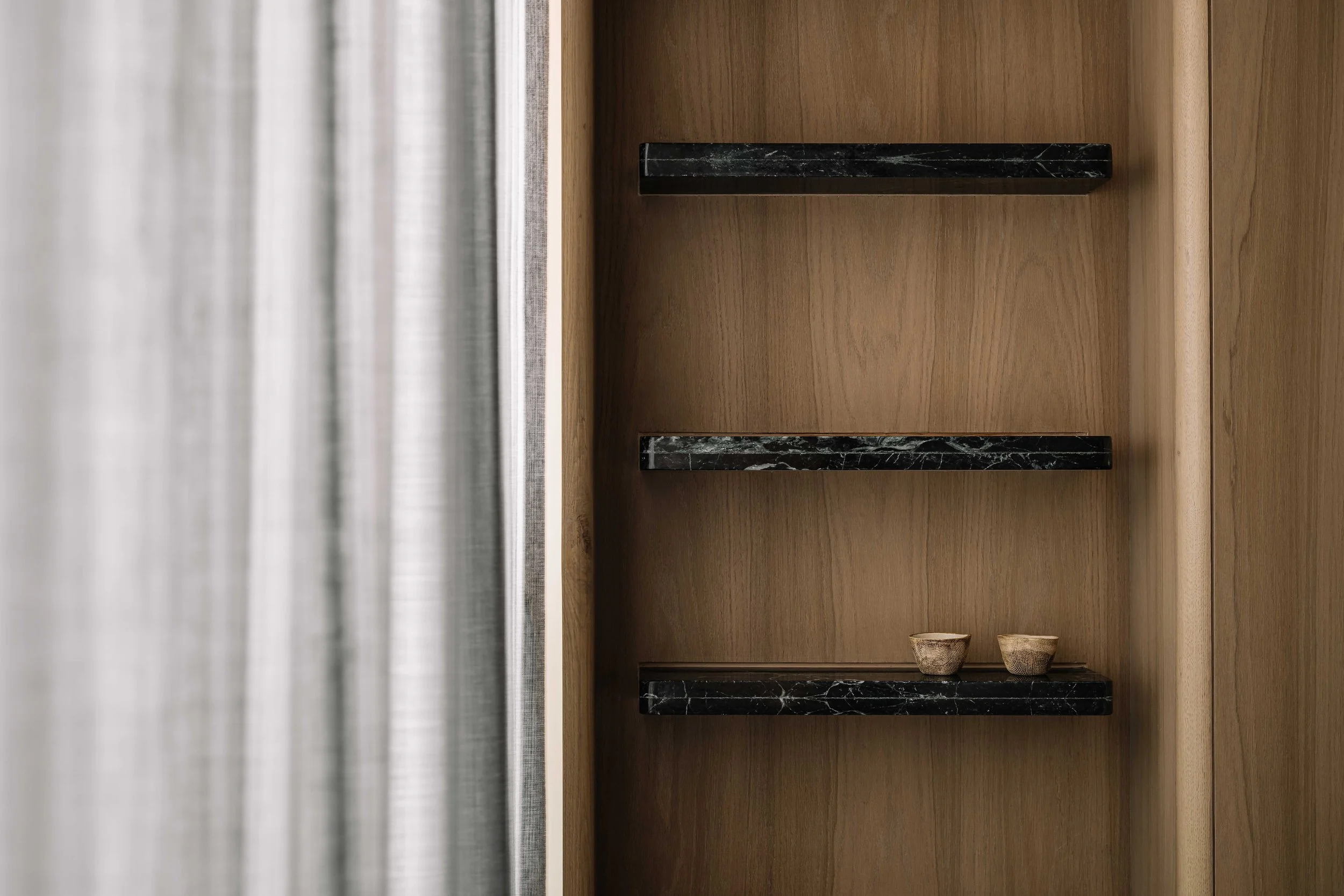 Wooden shelf unit with three black marble shelves and two small pottery cups on the bottom shelf, next to a window with sheer curtains.