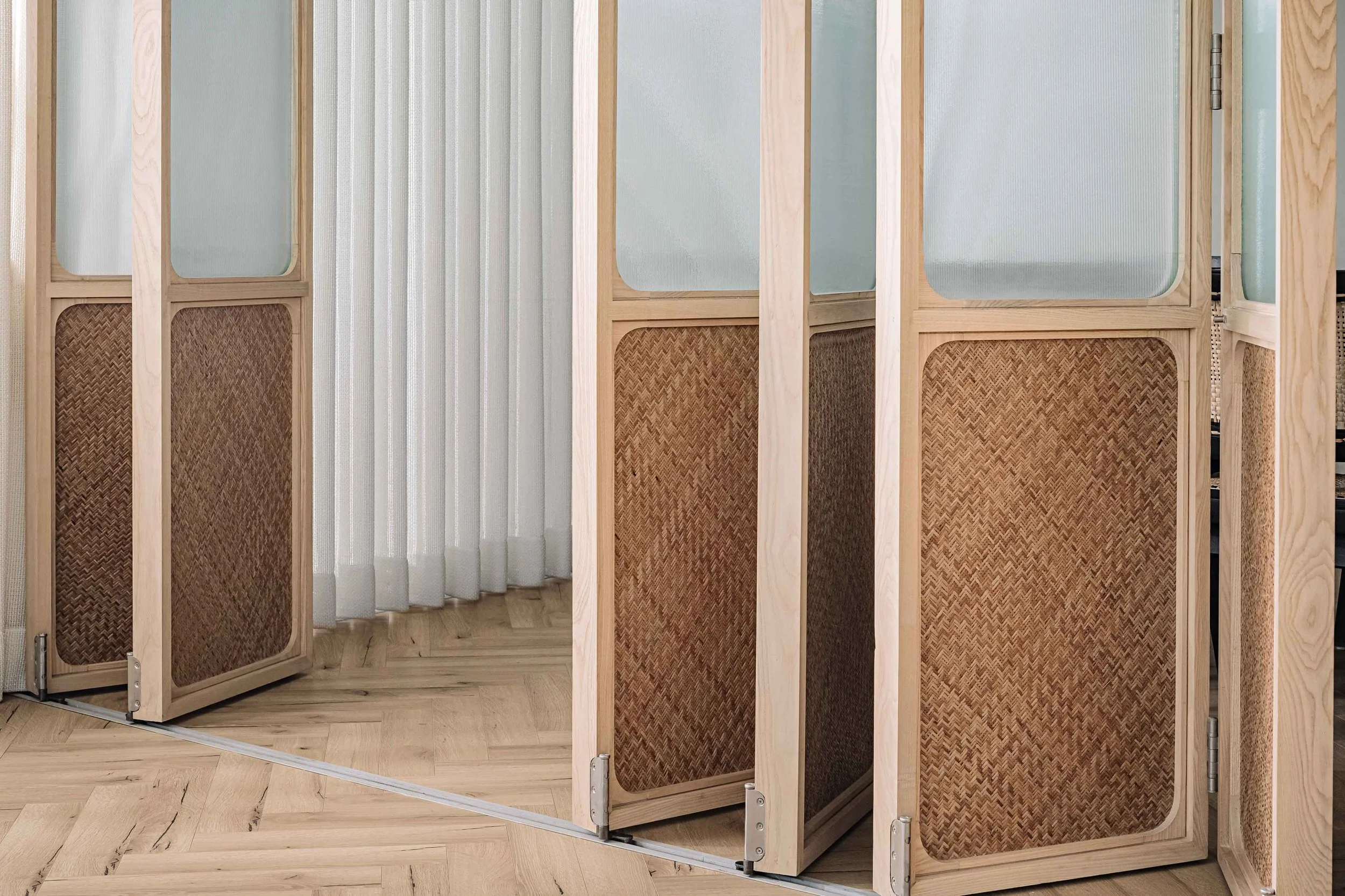 Wooden room divider with woven panels, set on a wooden floor with white curtains in the background.