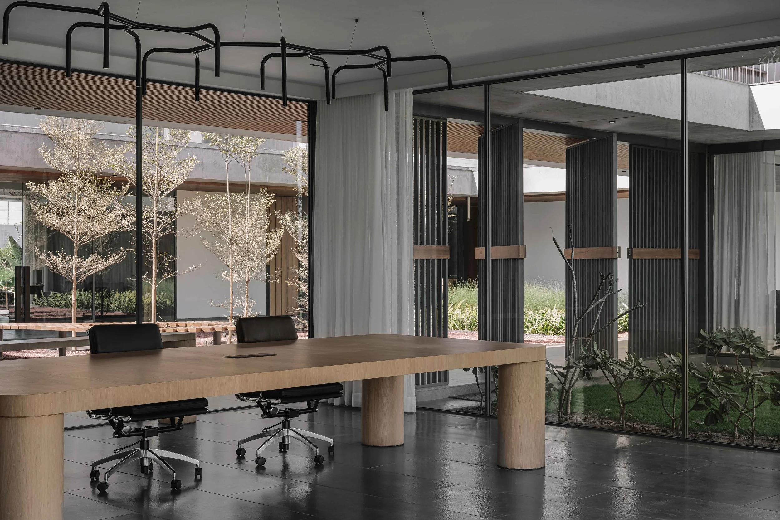 Modern conference room with a large wooden table, black chairs, glass walls, and indoor plants outside.