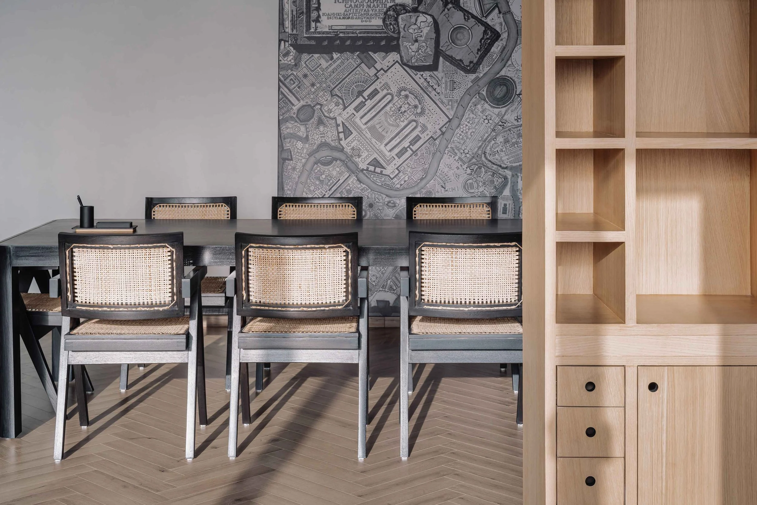 A dining room with a gray table surrounded by six chairs with black frames and beige woven rattan backs and seats. There is a dark-colored container with a straw and a closed book on the table. The wall behind the table features a large, detailed map