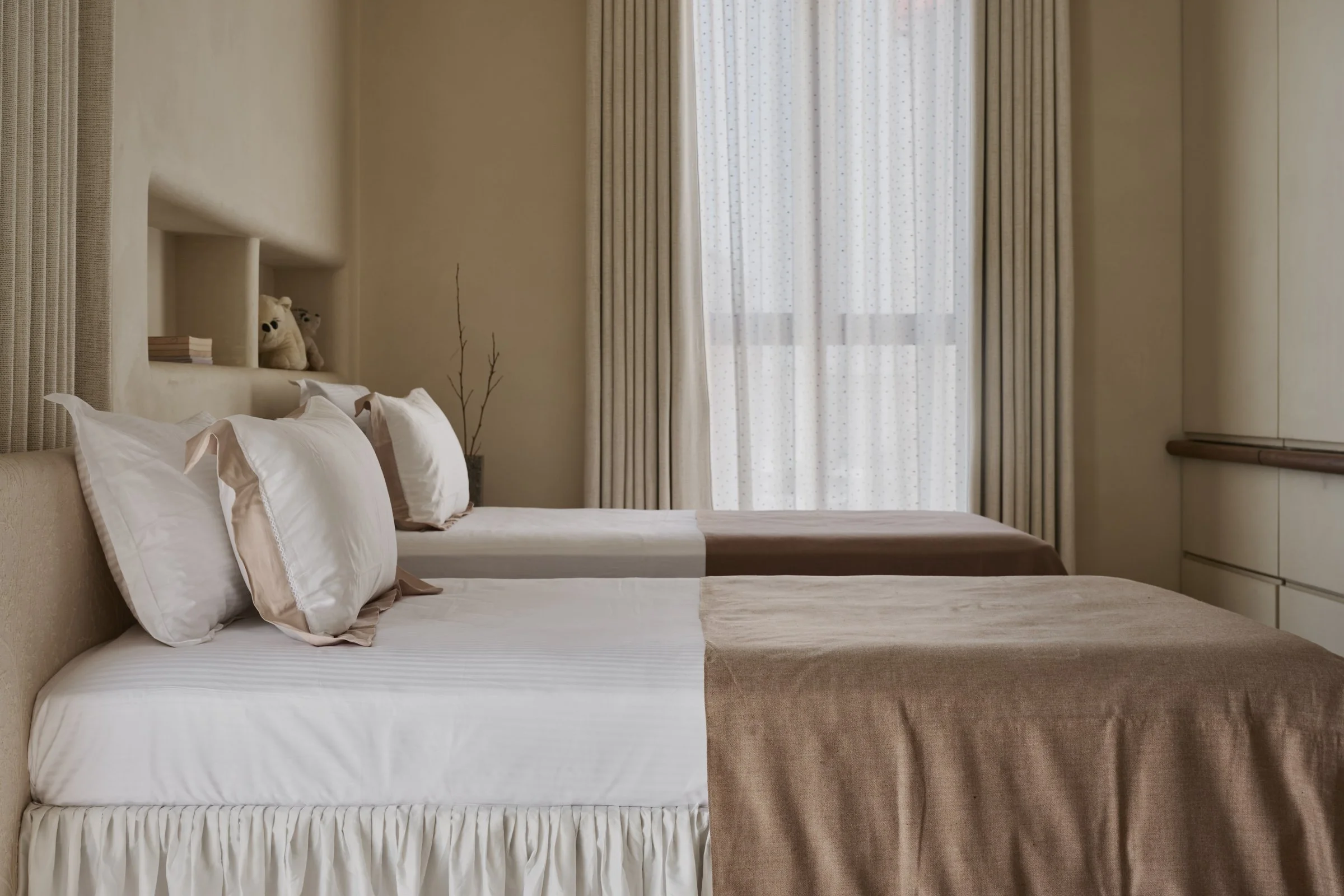Two neatly made beds with white pillows and beige bedspreads, positioned side by side in a hotel room, with a window covered by sheer and curtain drapes in the background.