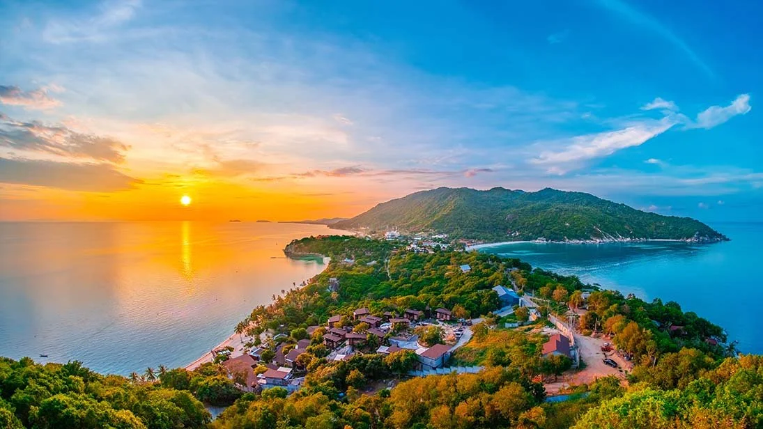 Blick auf eine Bucht bei Sonnenuntergang, umgeben von grünen Hügeln und einer kleinen Siedlung