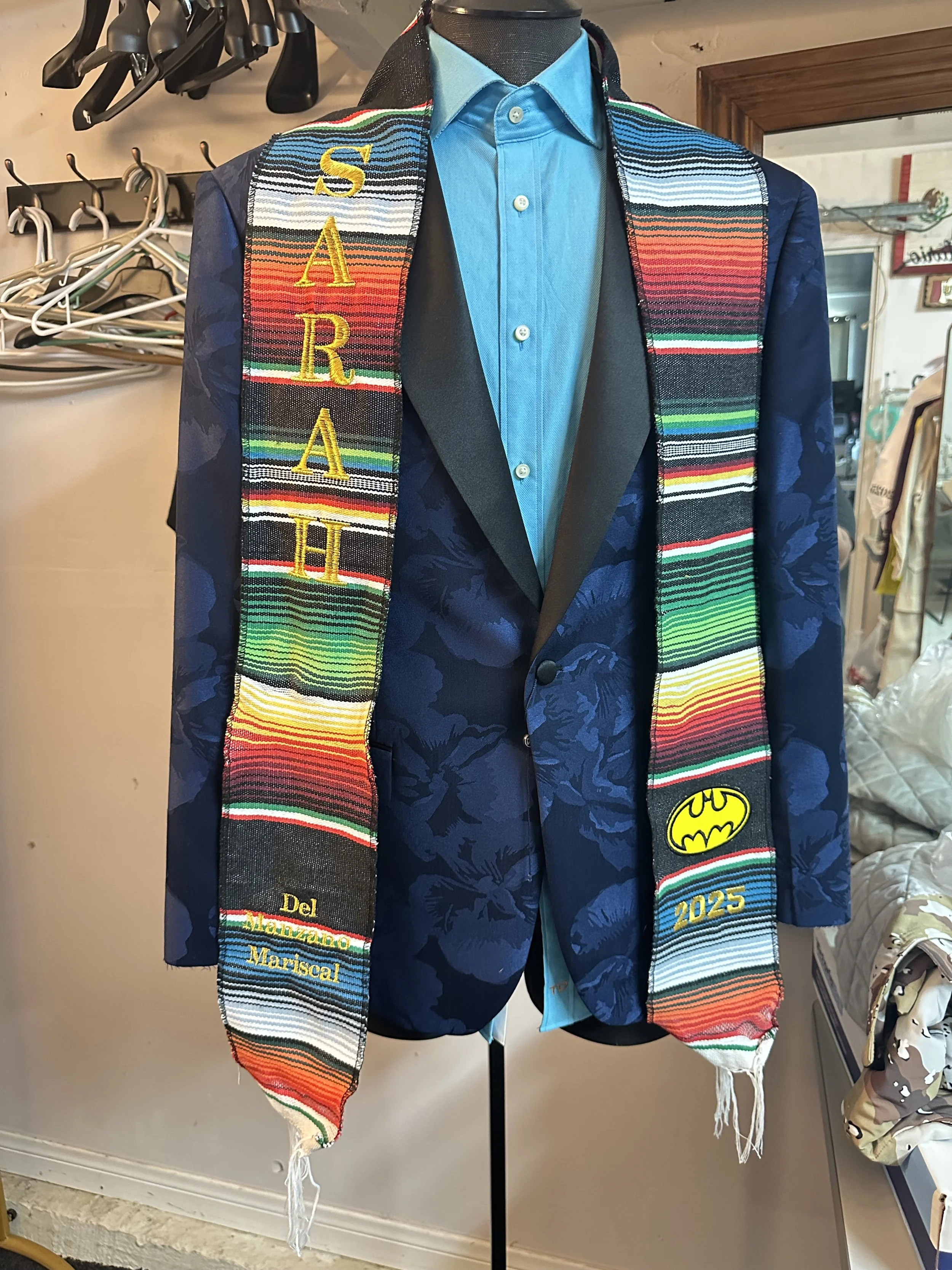 A mannequin dressed in a blue floral suit jacket, a bright blue button-up shirt, and a colorful, striped graduation stole with embroidered text and symbols, including the Batman logo.
