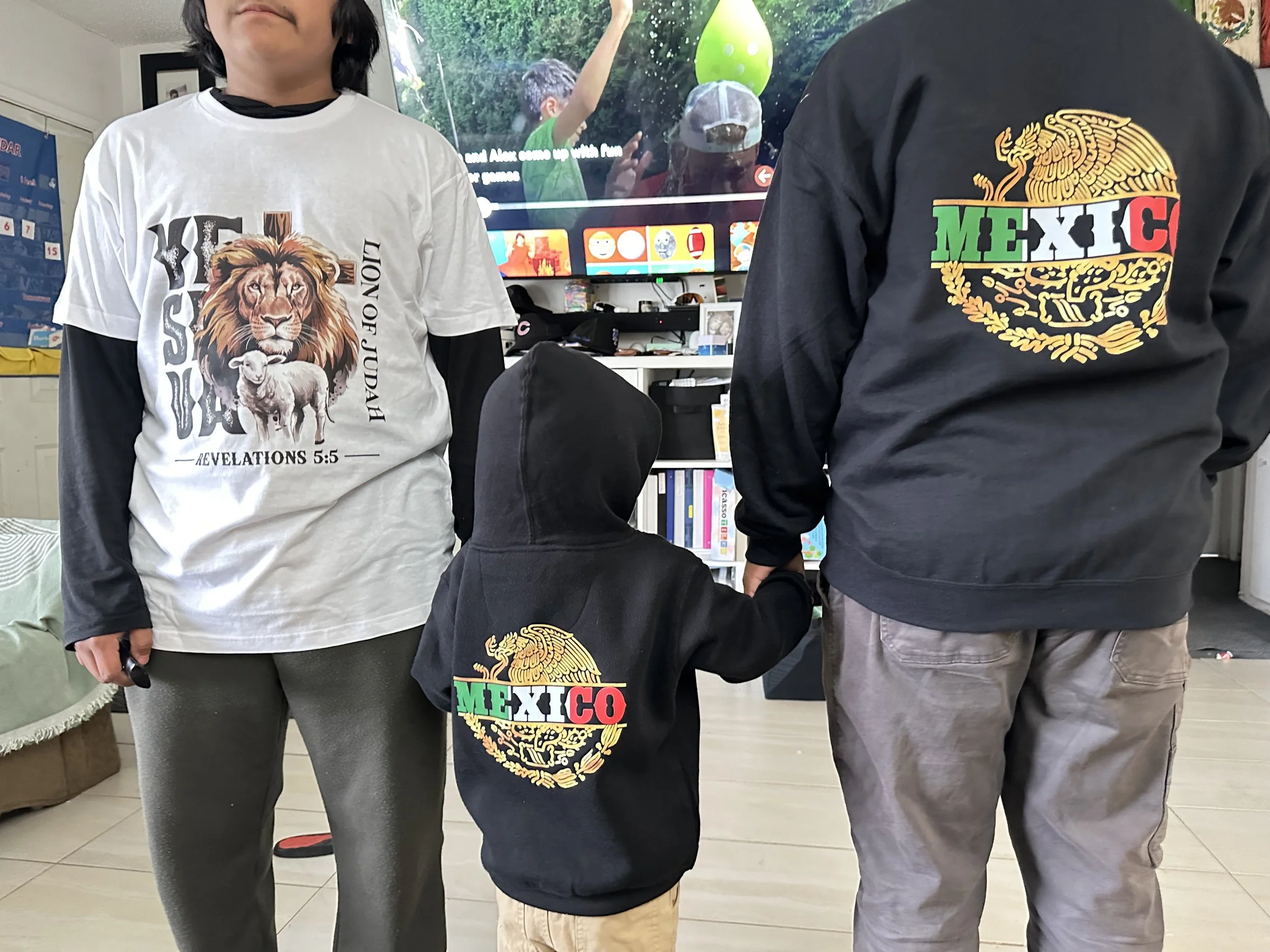 Three children, two adults, standing in a room with a TV and bookshelves, holding hands. The children are wearing hoodies with Mexican pride symbols, the girl with a white t-shirt that has a lion, sheep, and a Bible verse, the boy with a black hoodie