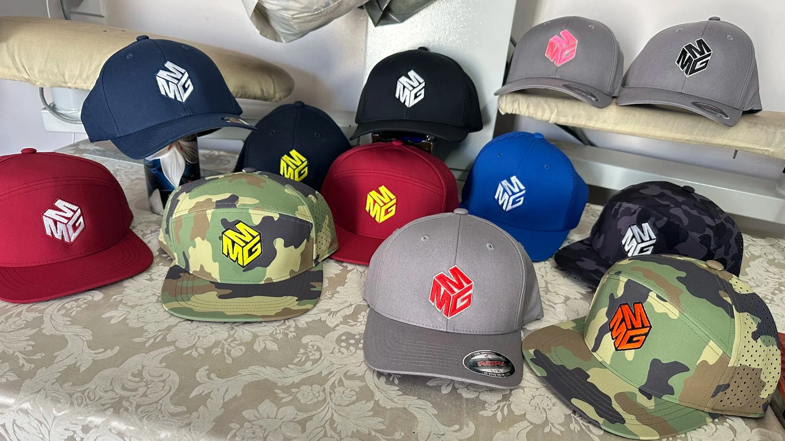 Various baseball caps with different colors and patterns arranged on a table, featuring a logo of a stylized 3D letter M in different colors.