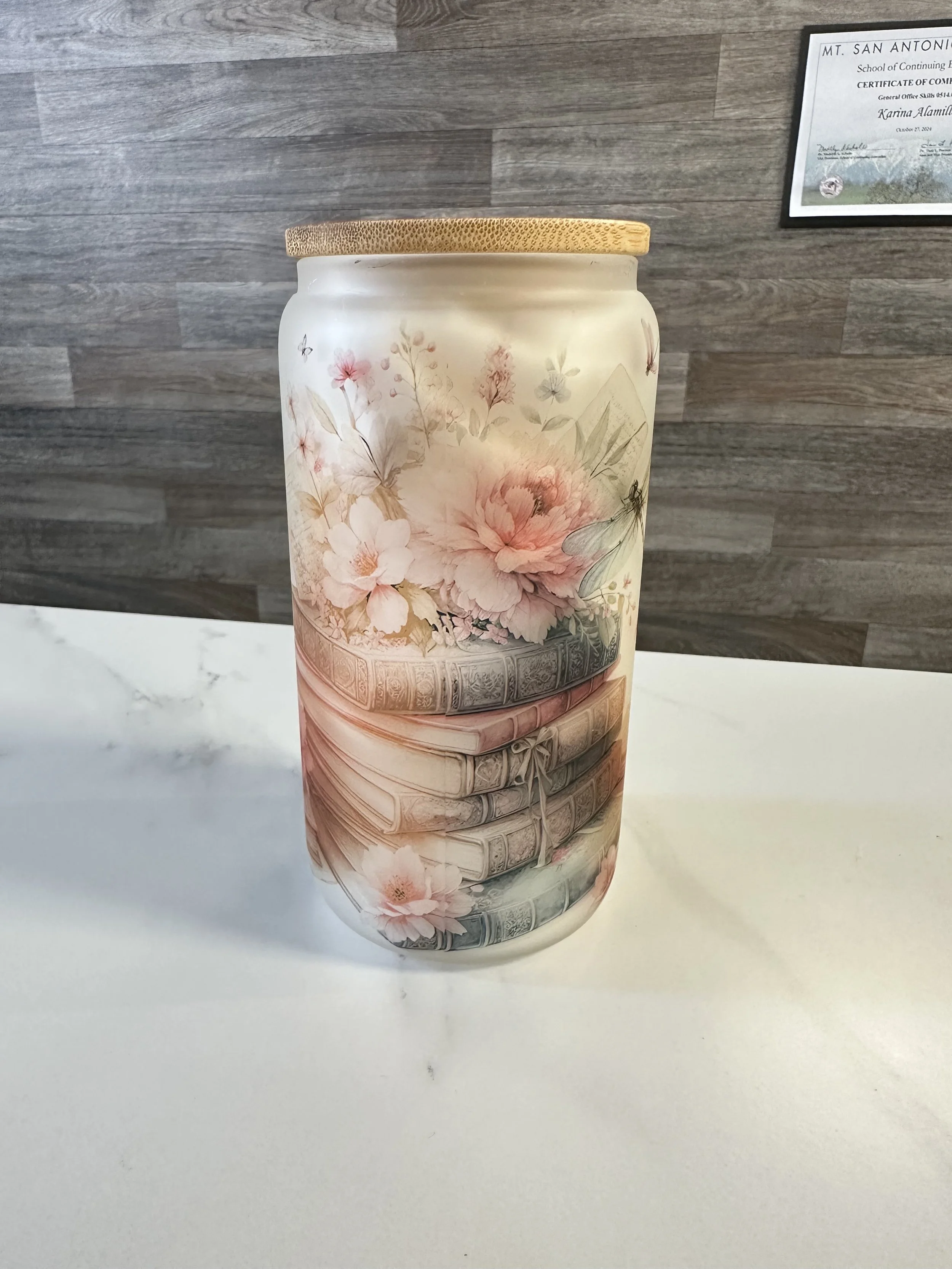 Decorative jar with a floral and book stack design on a white surface, with a wooden wall and a framed certificate in the background.