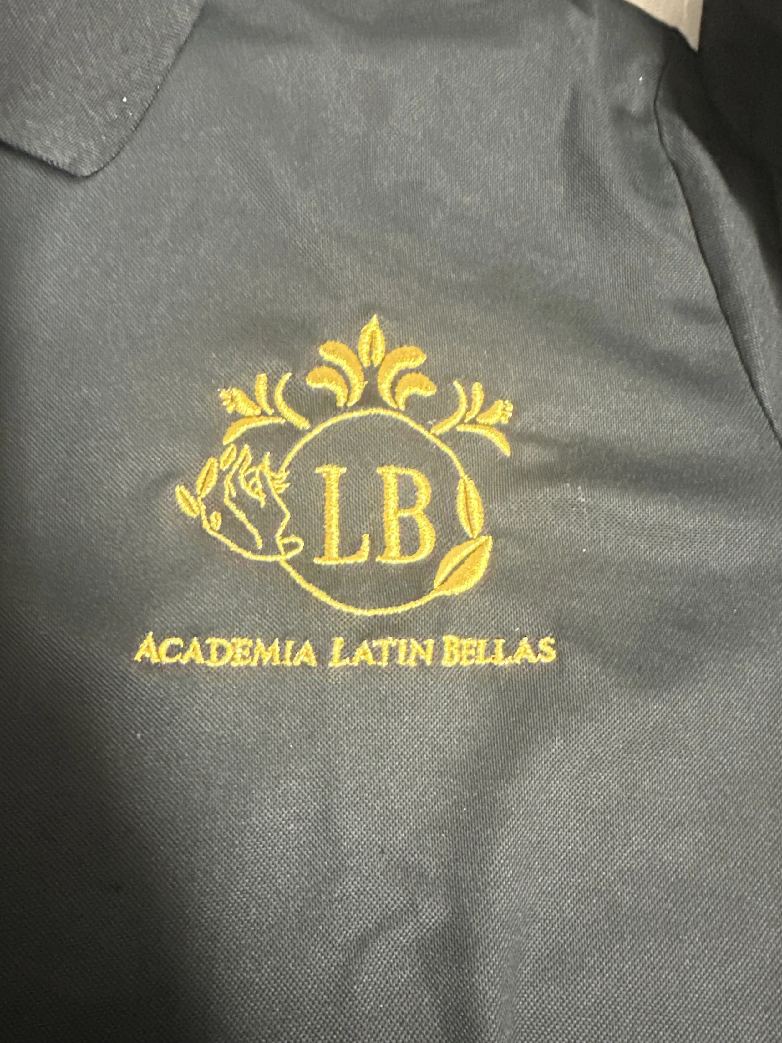 Close-up of a black fabric with yellow embroidered logo and text that reads 'LB Academia Latin Bellas'.