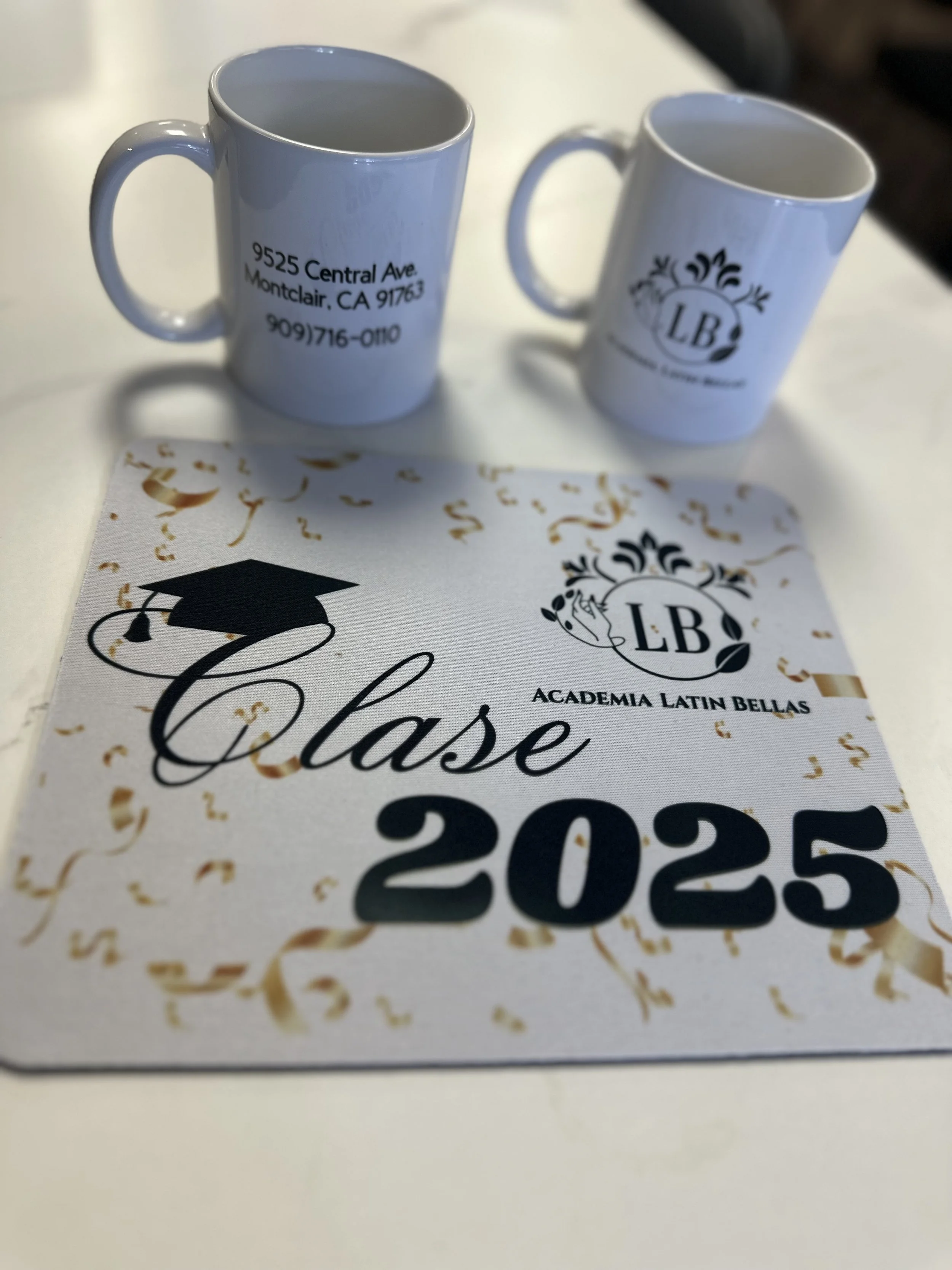 Two white coffee mugs with printed text and logos, one showing an address in Montclair, California, and the other with a logo and initials 'LB'. A printed program or certificate with the text "Clase 2025," a graduation cap illustration, and the logo