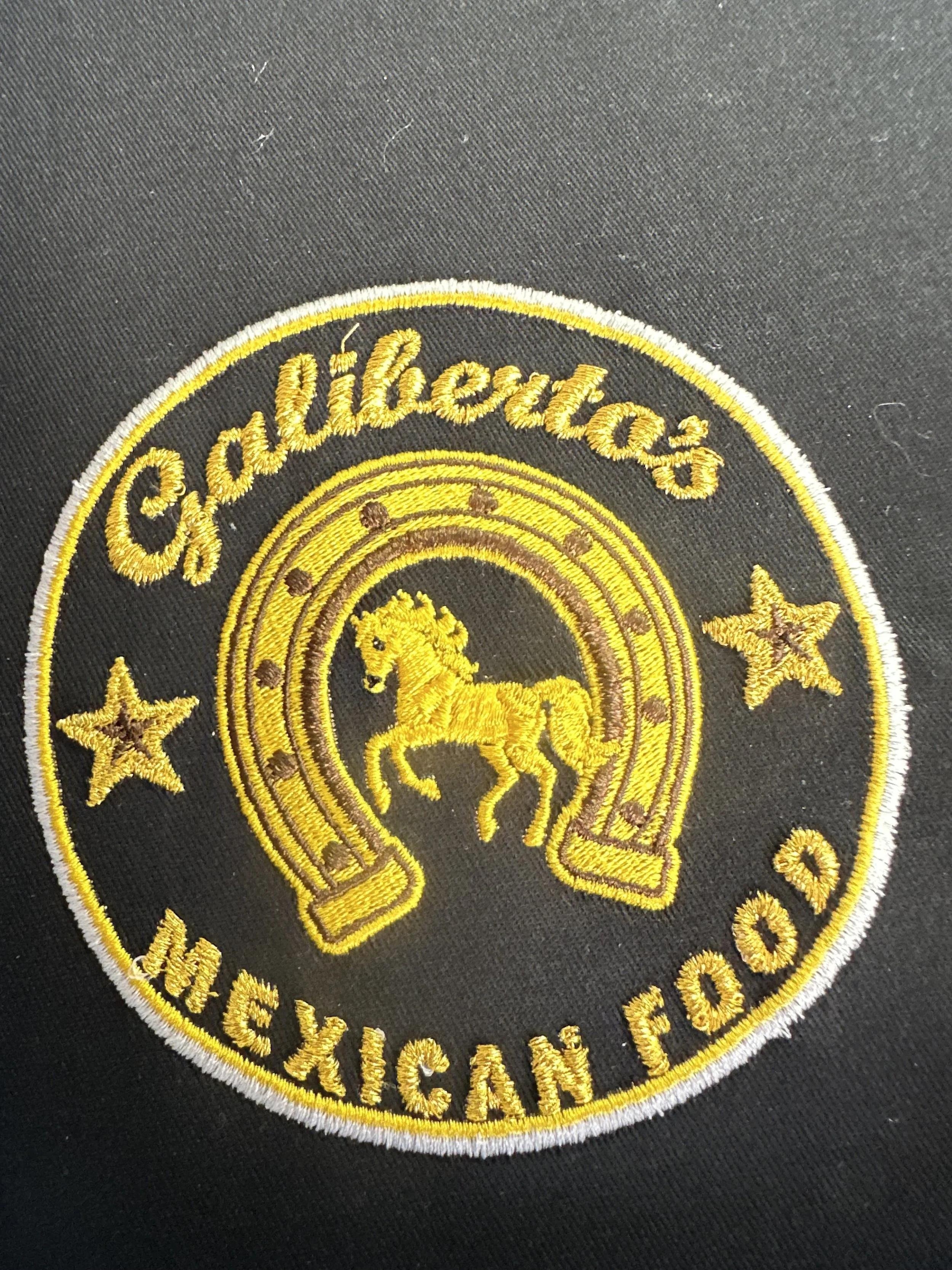 A circular embroidered patch with gold and yellow design, featuring the words 'Gallibertaz Mexican Food', a horse inside a horseshoe, and two stars on each side.