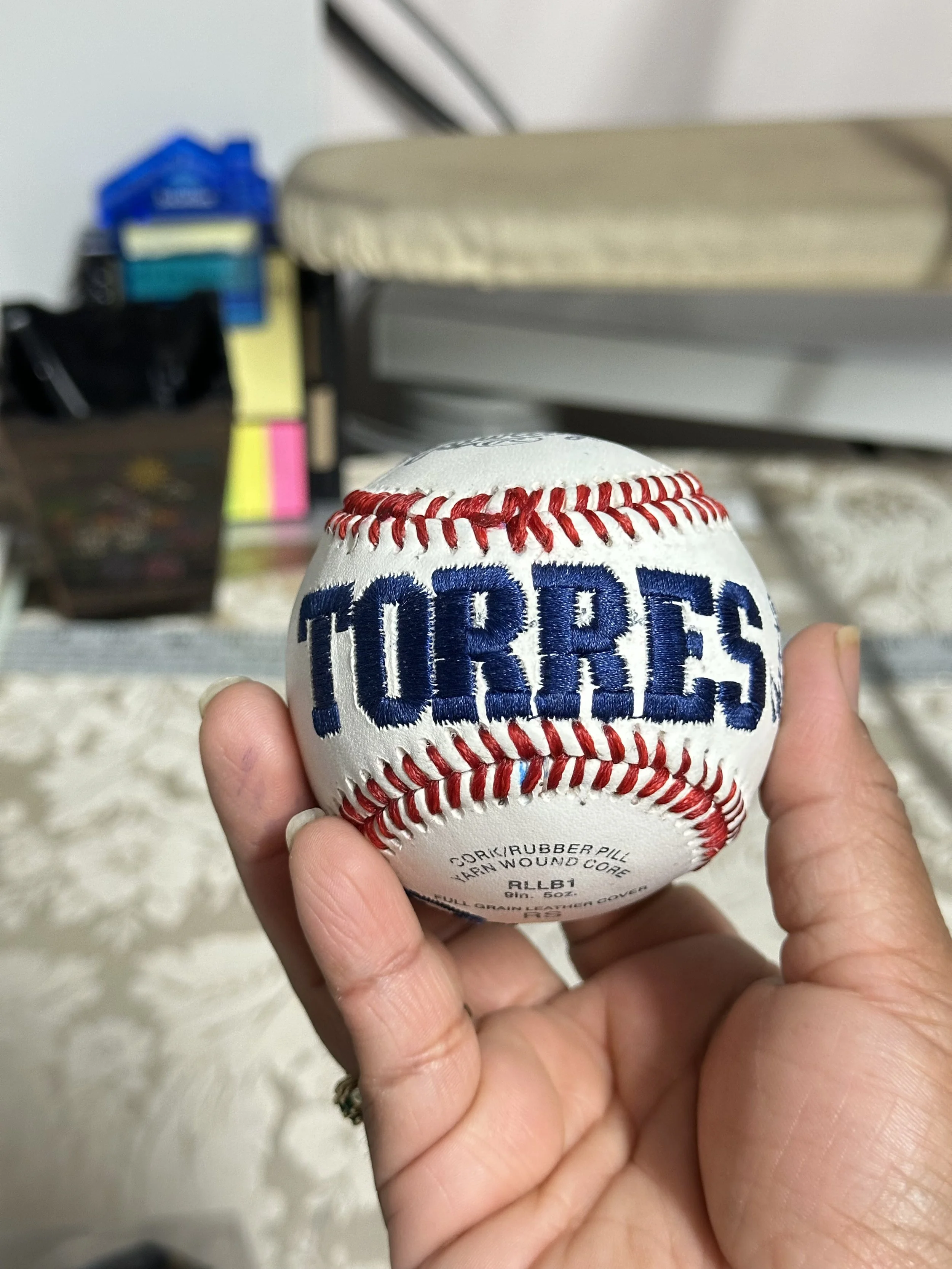 A hand holding a white baseball embroidered with the word 'TORRES' in blue. The background shows a cluttered room with various items, including a beige cushioned seat and some storage boxes.