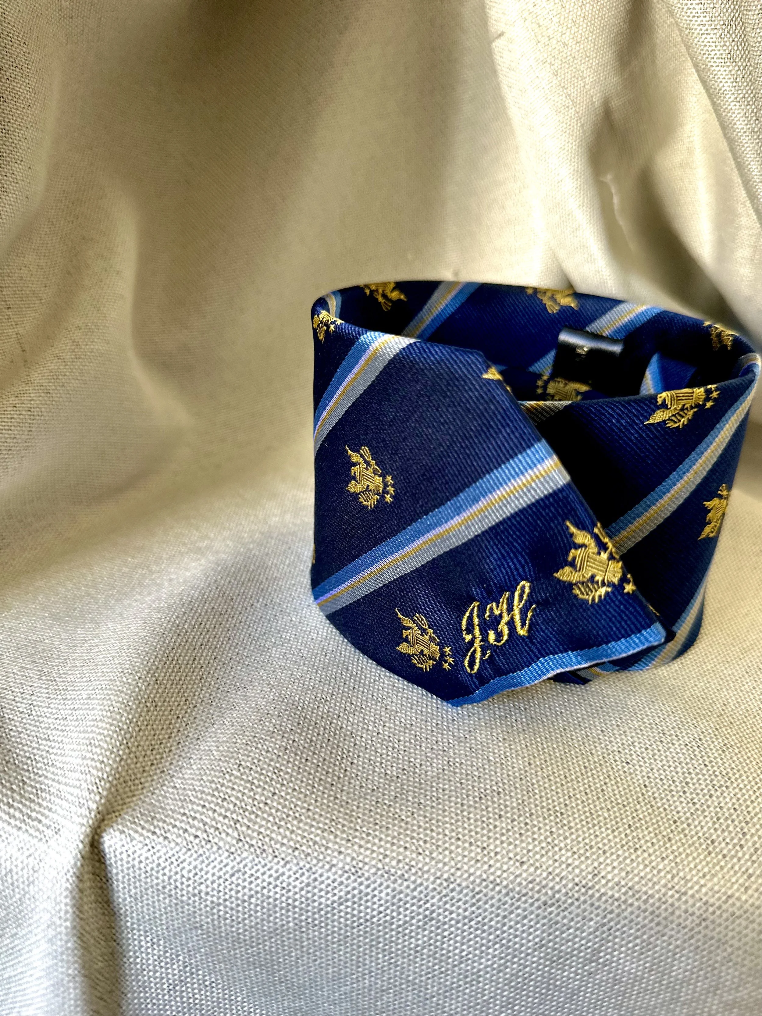 A blue and gold striped tie with initials, resting on a cream-colored fabric background.