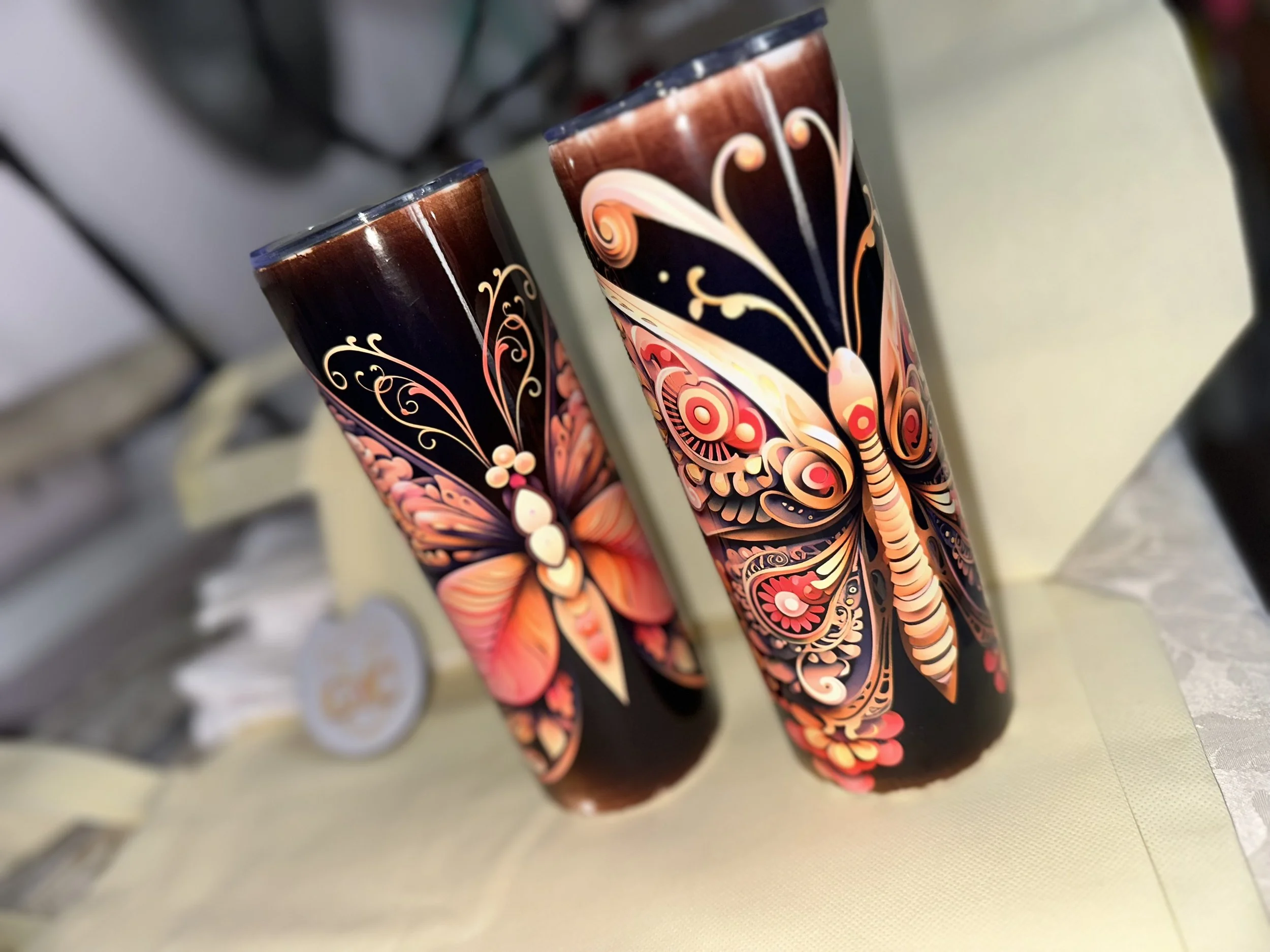 Two tall black tumblers with colorful butterfly and floral designs in pink, orange, and beige, placed on a table with a cream tablecloth.