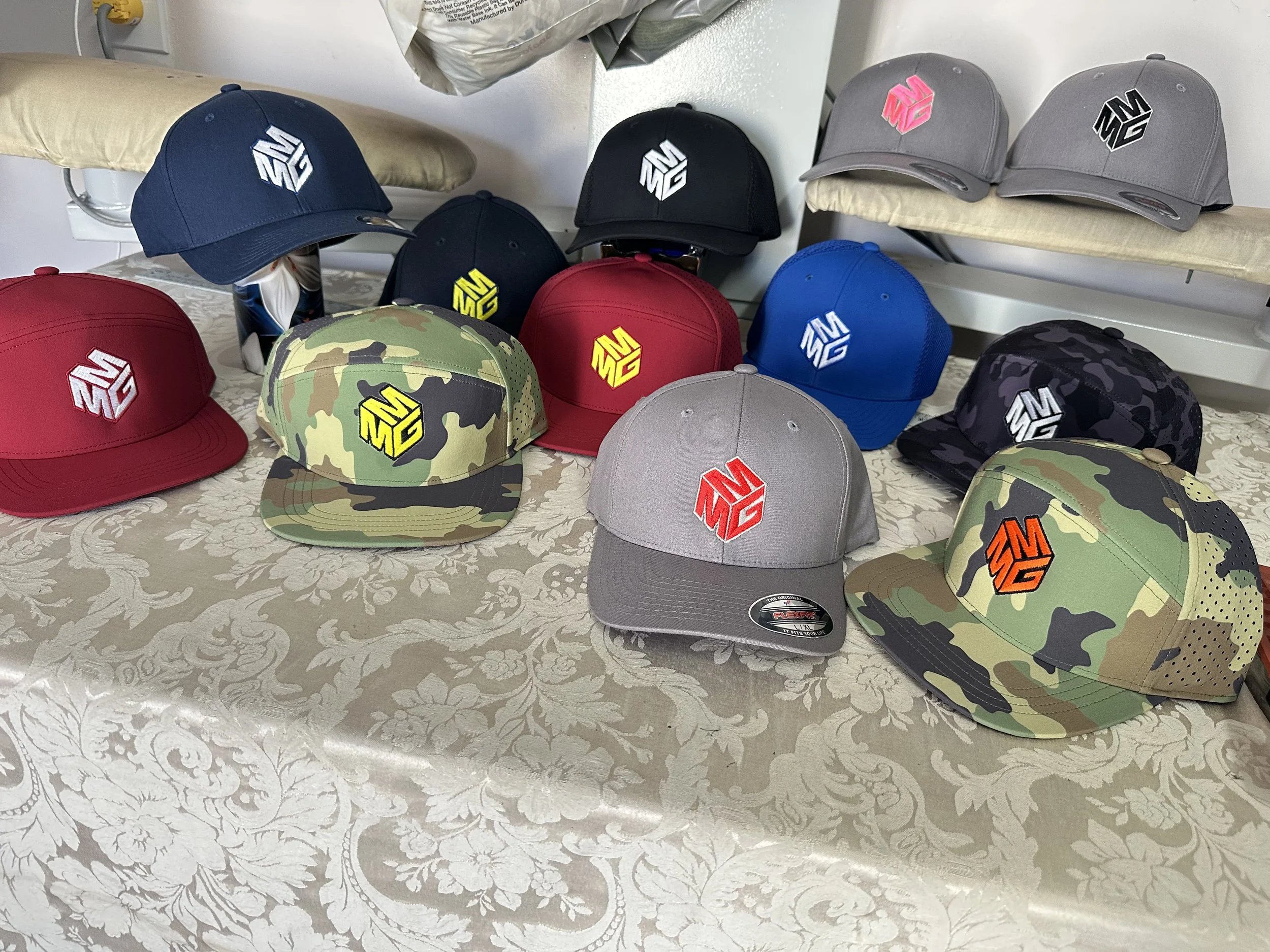Various baseball caps with different colors and patterns arranged on a table, featuring a logo of a stylized 3D letter M in different colors.
