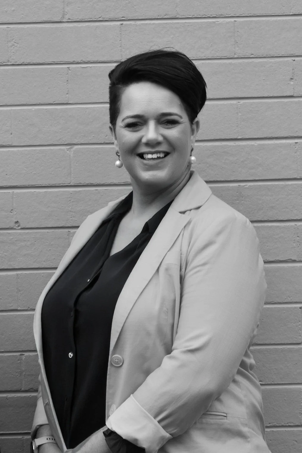 Black and white photo of a woman with short dark hair, earrings, wearing a light-colored blazer and a dark shirt, smiling in front of a brick wall.