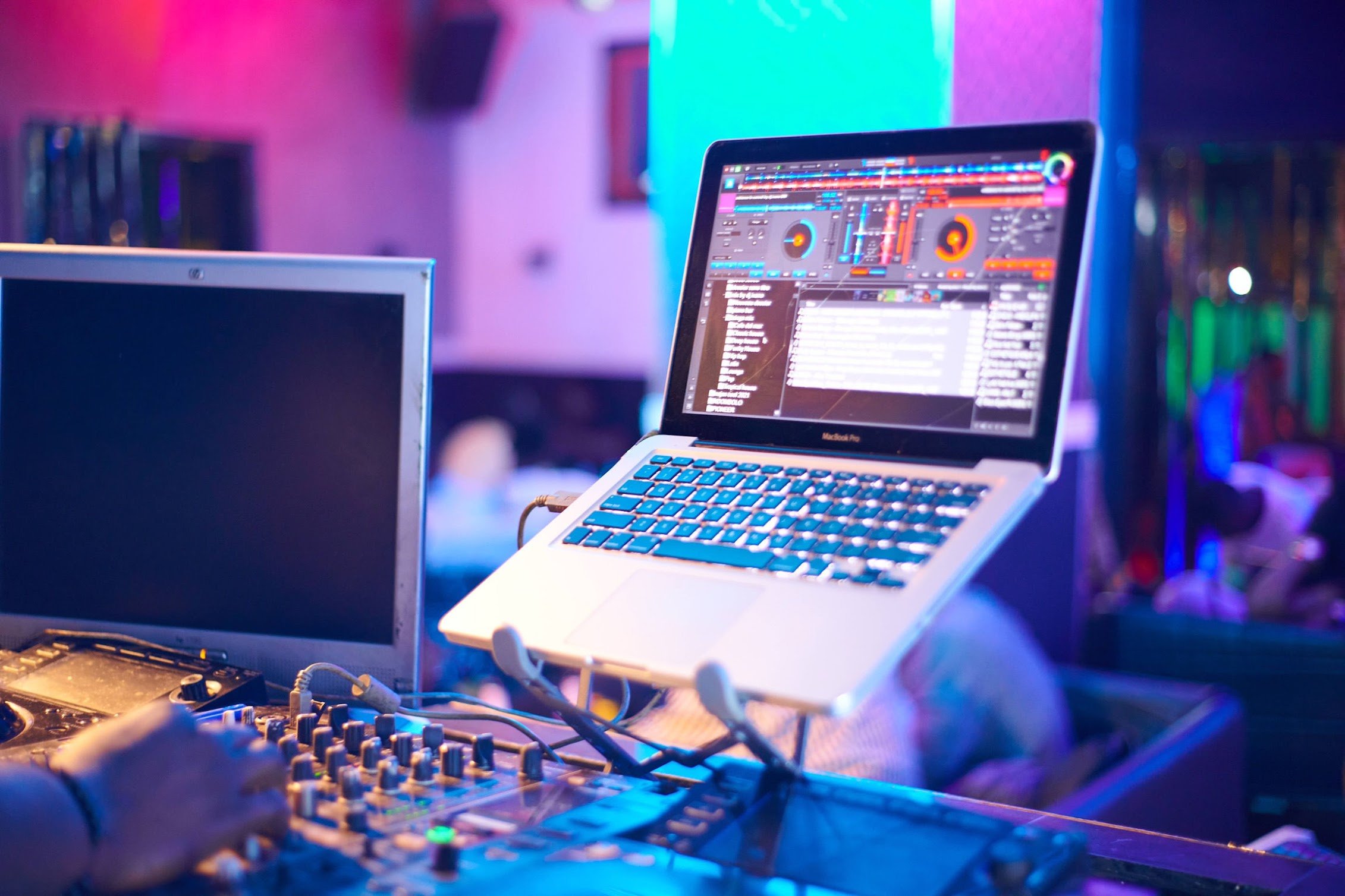A DJ setup featuring a laptop displaying a digital music mixing software, with a DJ mixer and a monitor in a colorful, dimly lit club or party environment.