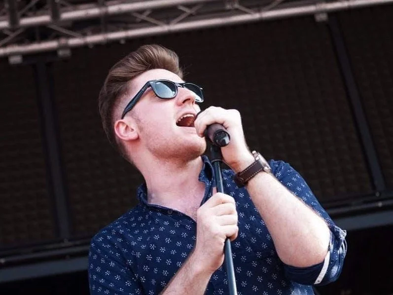 Young man with sunglasses singing into a microphone on stage.
