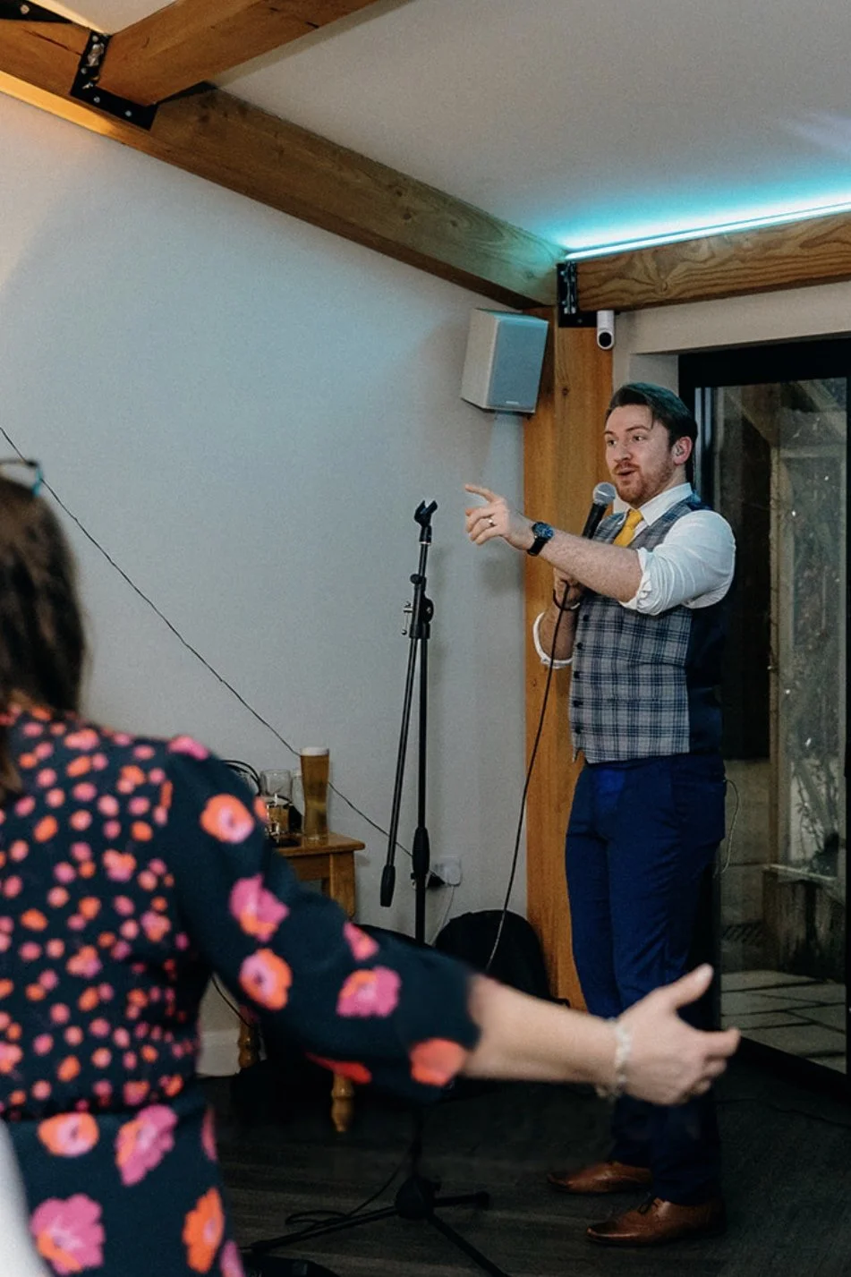A man in formal attire, holding a microphone, speaking at an indoor event. A woman in a floral dress is visible in the foreground, gesturing with her hand. The setting appears to be a decorated room with wooden beams and a speaker mounted on the wall.