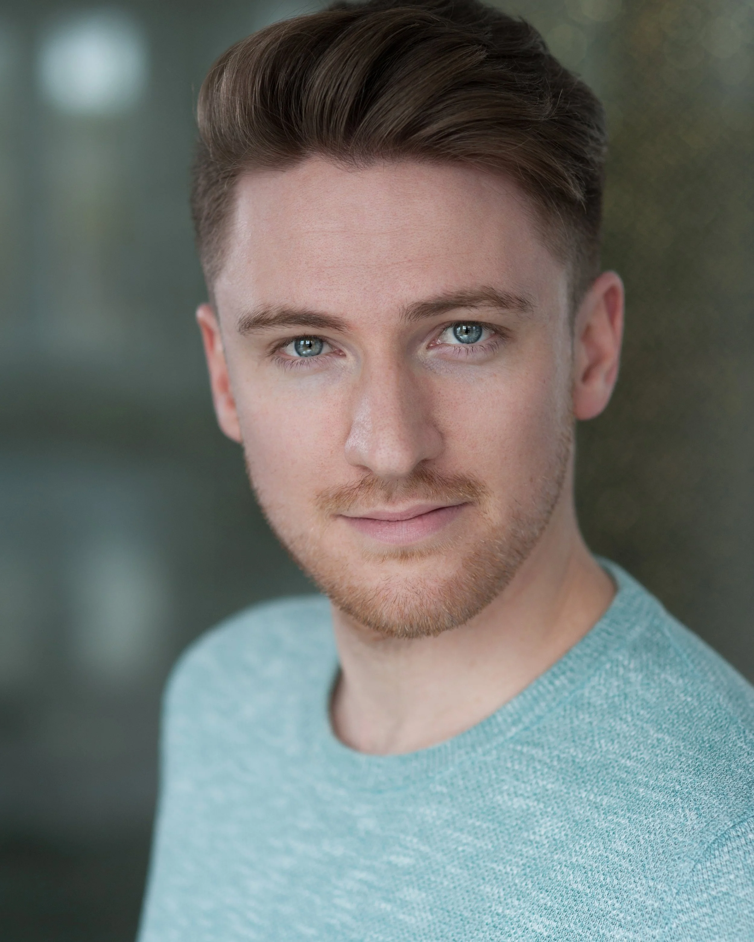 Close-up portrait of a young man with blue eyes, light brown hair, and a light beard, smiling slightly and looking into the camera, wearing a light blue shirt.
