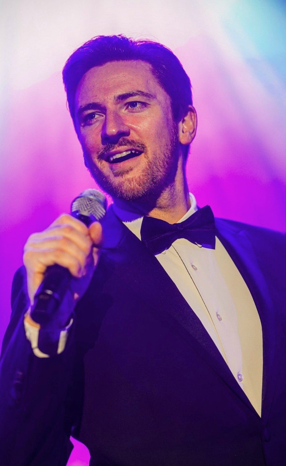 Man in tuxedo holding a microphone on a stage with colorful purple and blue lighting