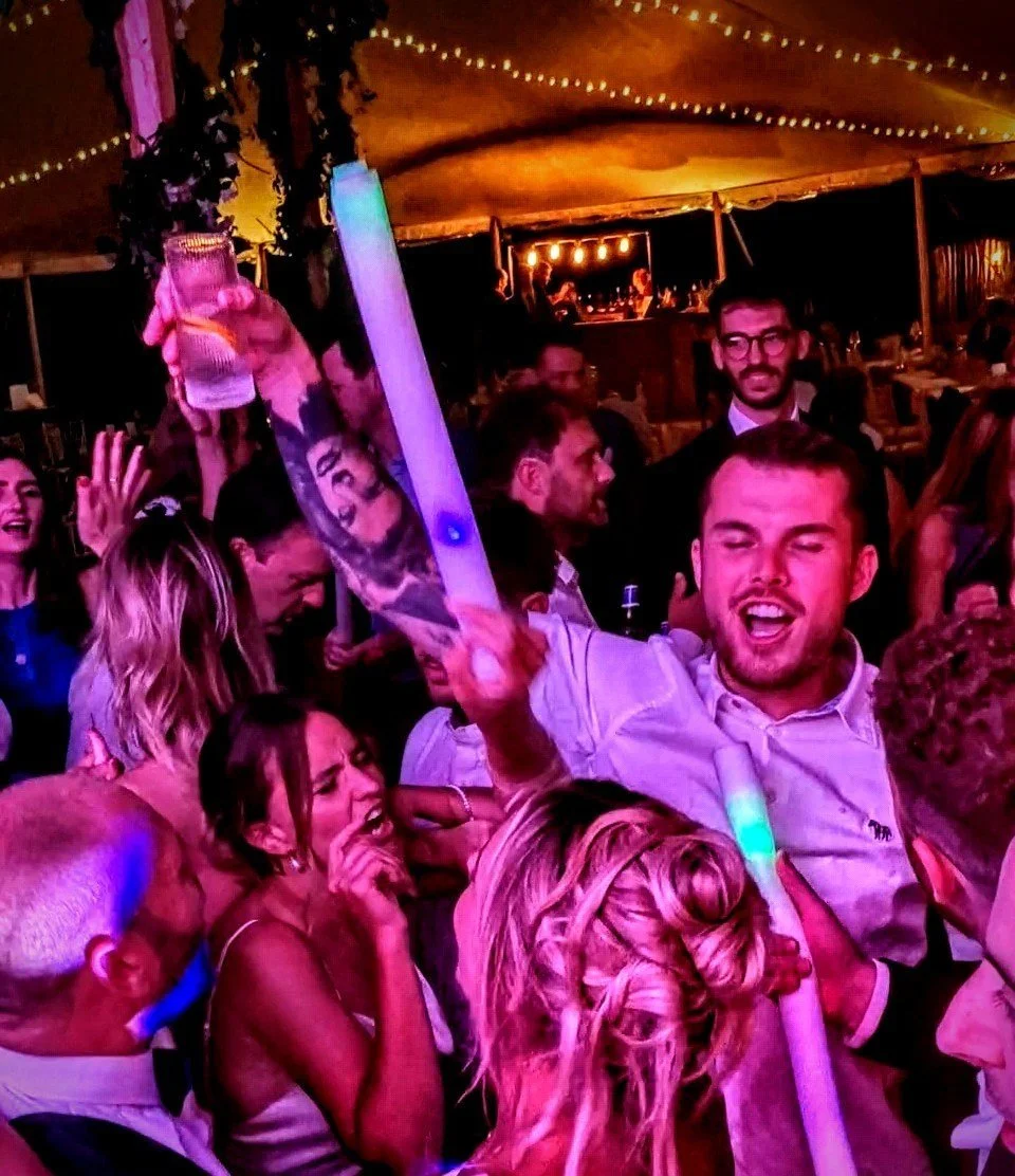 People celebrating at a lively party, some holding glow sticks and drinks, with cheerful expressions under warm, colorful lighting.