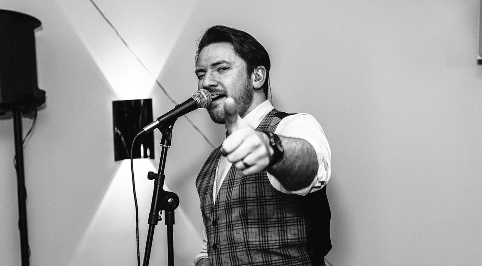 A man with dark hair and a beard, wearing a vest over a white shirt, pointing at the camera while singing into a microphone.