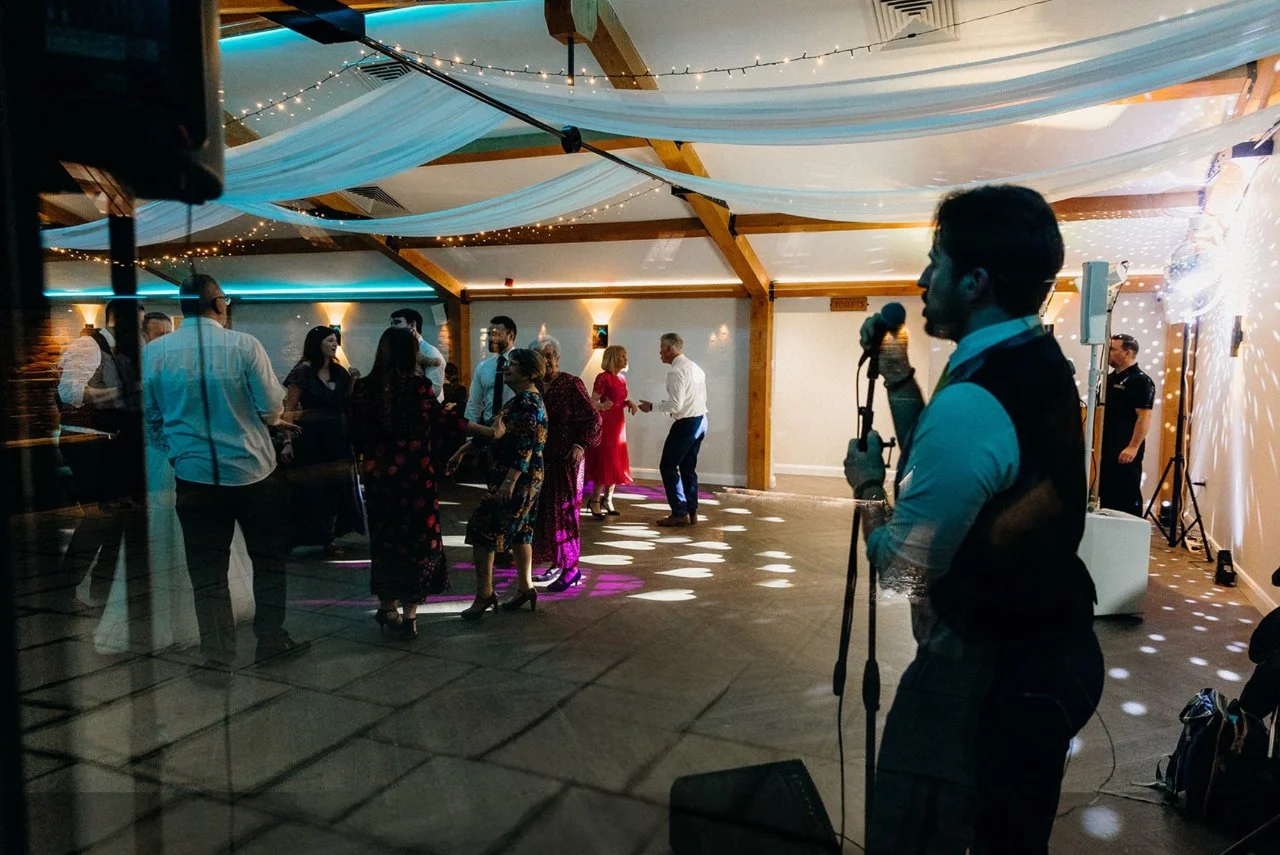 People dancing and socializing at a celebration or wedding with a singer performing in the foreground. Decor includes string lights and draped fabric on the ceiling.