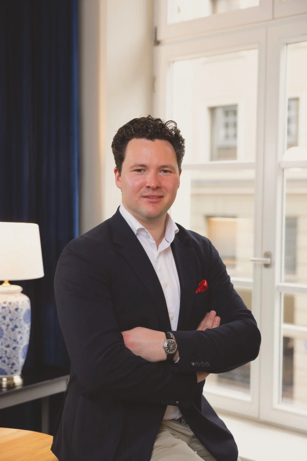 Ein Mann in dunklem Anzug mit weißen Hemd und roter Einstecktuch steht in einem hell erleuchteten Raum mit großen Fenstern. Er hat kurze, lockige Haare und trägt eine Armbanduhr.