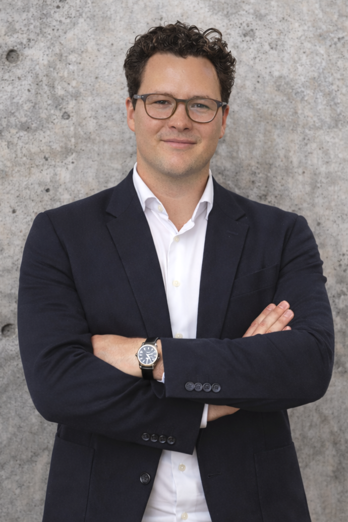 Ein Mann trägt eine Brille, einen weißen Hemd und einen schwarzen Blazer, steht vor einer Betonwand, und hat die Arme verschränkt.