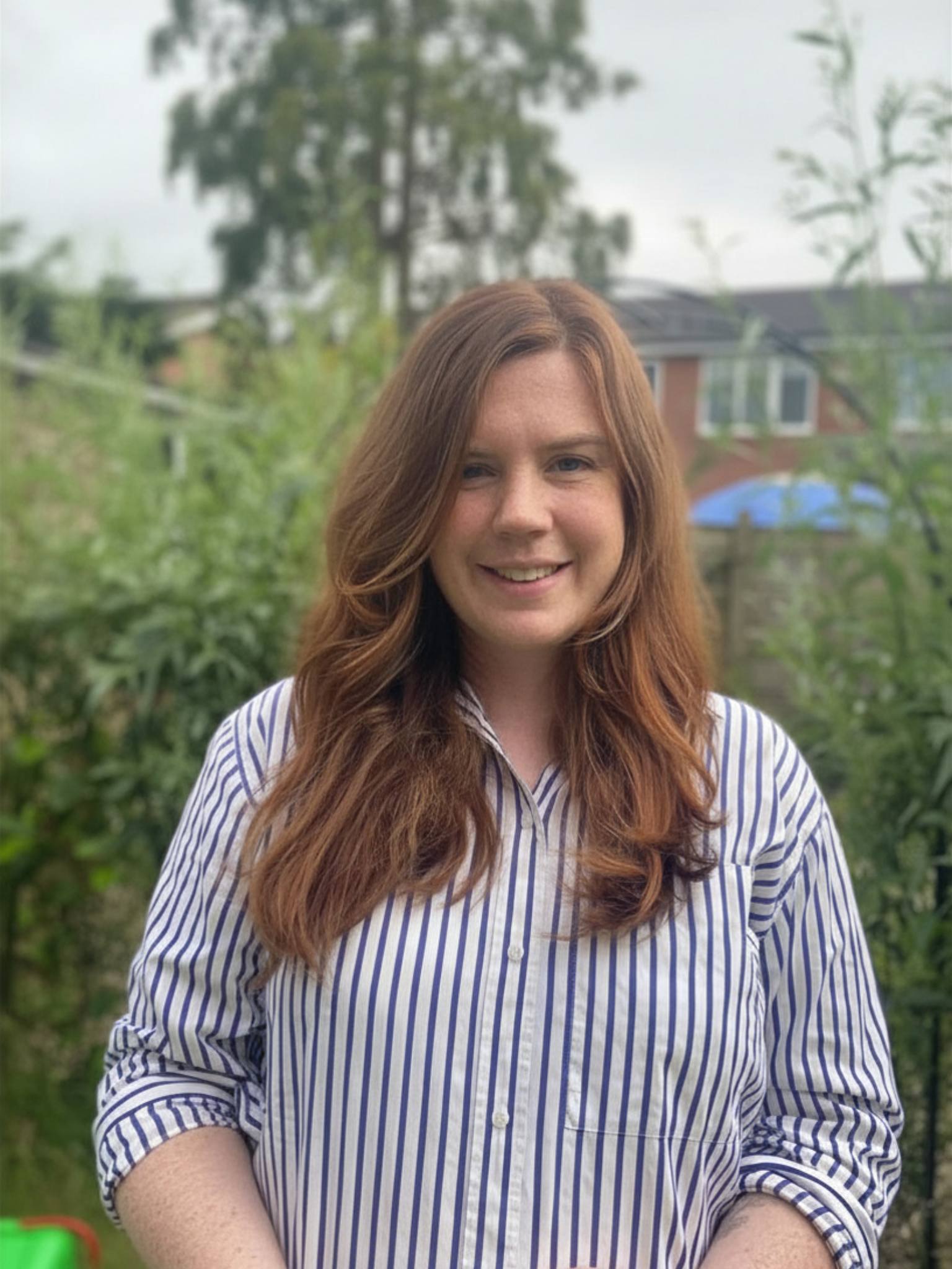 A woman with long red hair wearing a blue and white striped shirt standing outdoors with greenery and trees in the background.